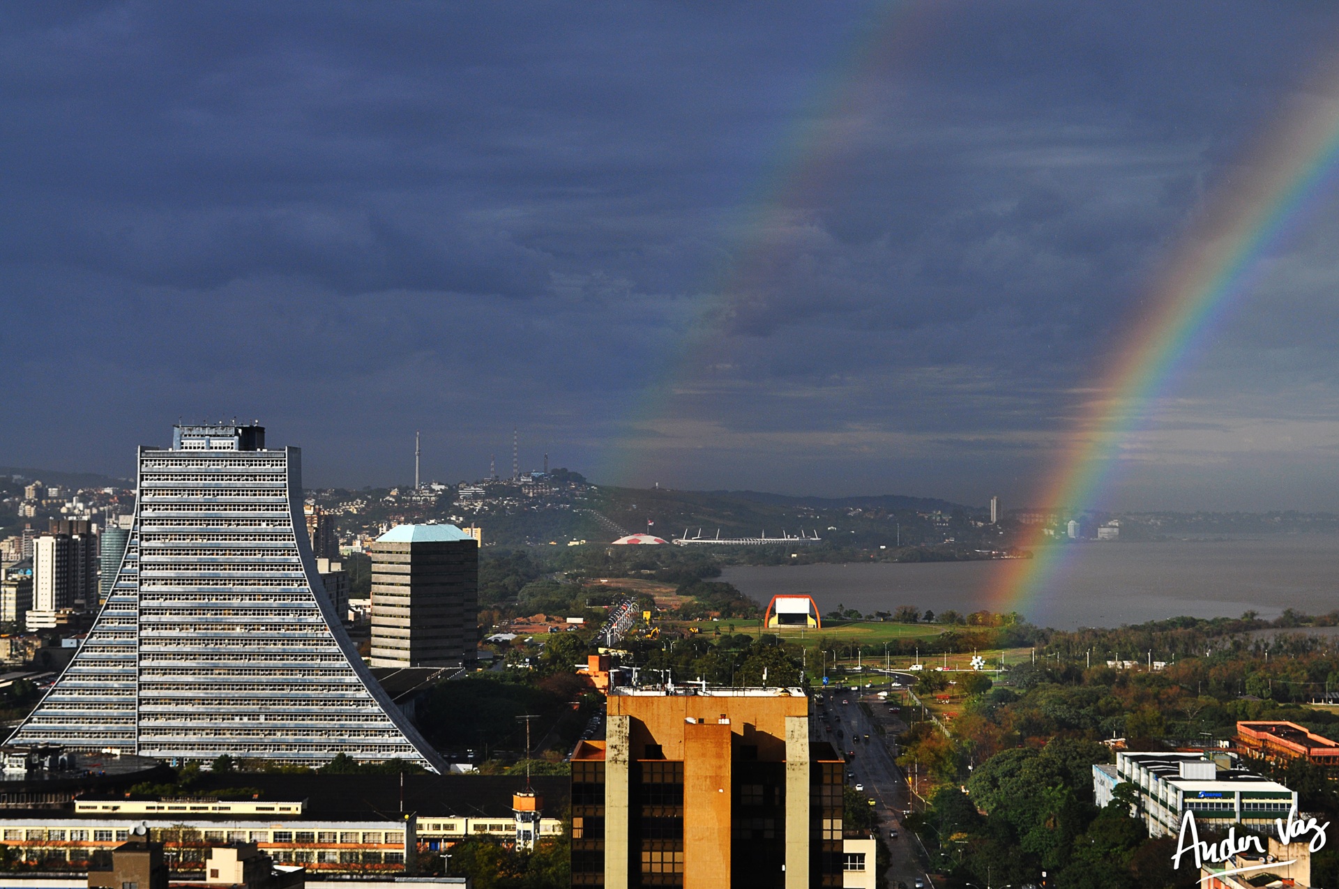Vista panorâmica de Porto Alegre com destaque para o edifício do Centro Administrativo Fernando Ferrari (CAFF), no bairro Praia de Belas. À esquerda, o prédio modernista em formato piramidal domina a paisagem urbana, enquanto ao fundo um arco-íris duplo colore o céu sobre o Guaíba