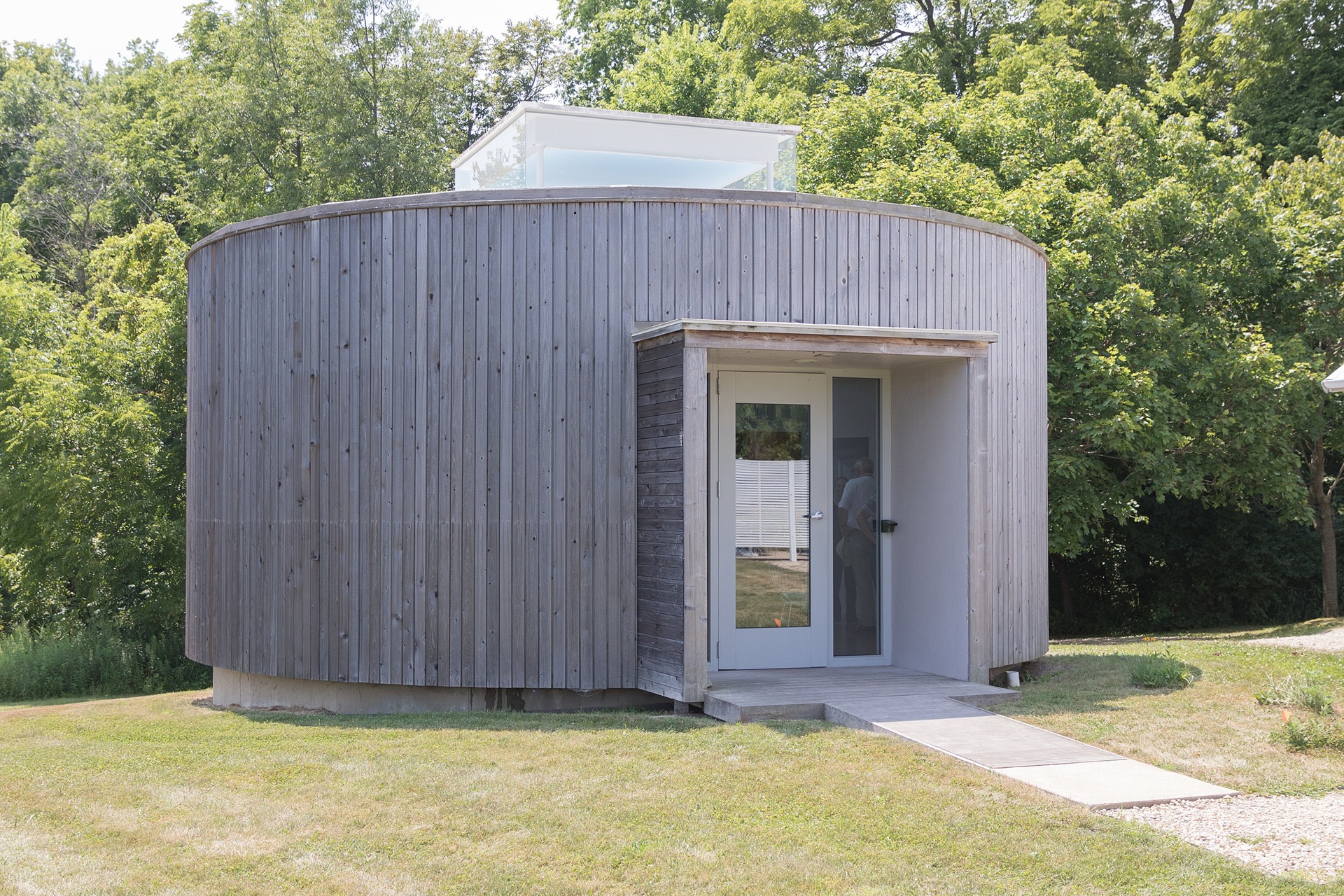 Vista externa da Galeria Barnsworth, estrutura circular de madeira com porta de vidro, localizada próxima à Farnsworth House