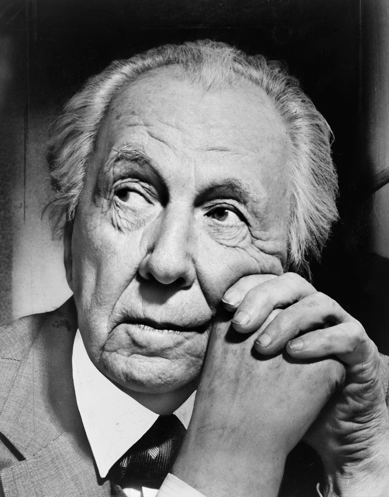 Fotografia em preto e branco de Frank Lloyd Wright. Na imagem, ele aparece com expressão reflexiva e as mãos entrelaçadas junto ao rosto