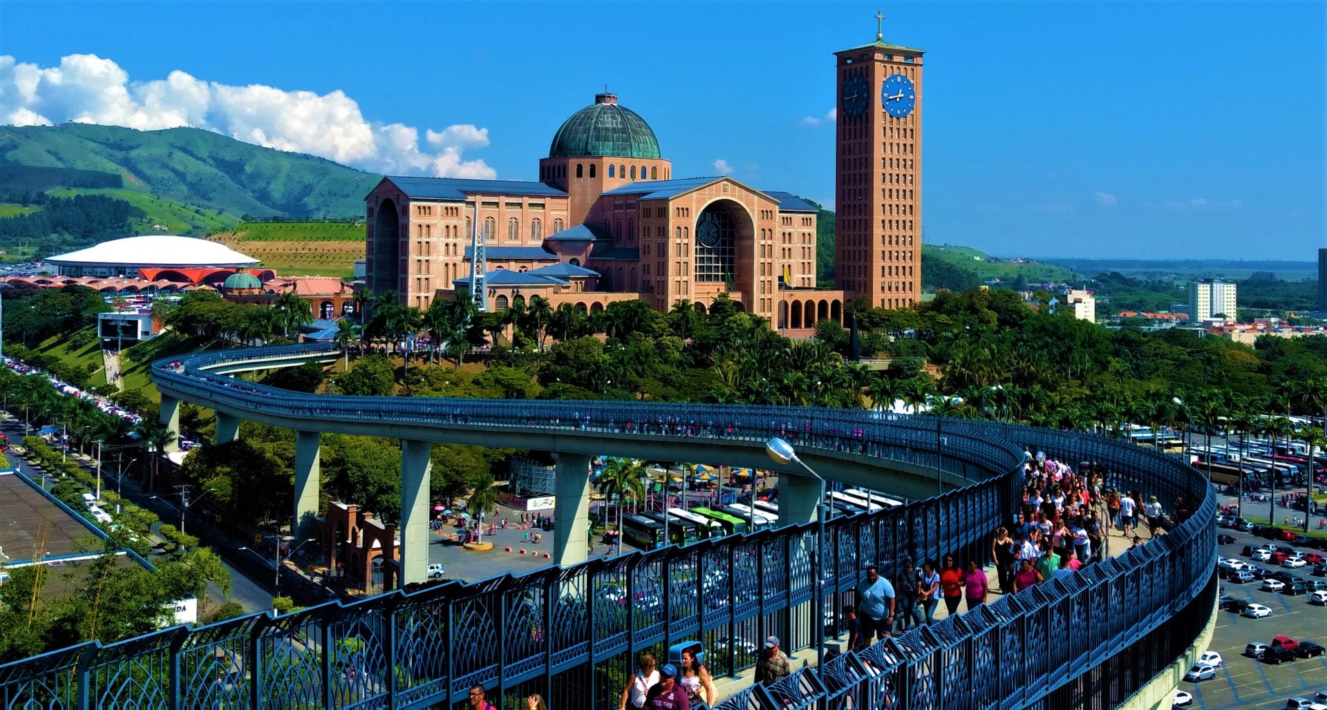 Vista aérea da Passarela da Fé, com a Basílica de Nossa Senhora Aparecida ao fundo. A passarela, em formato sinuoso, conecta a Basílica Velha ao Santuário Nacional, oferecendo uma visão panorâmica da cidade. O céu claro e a luz suave destacam os detalhes arquitetônicos