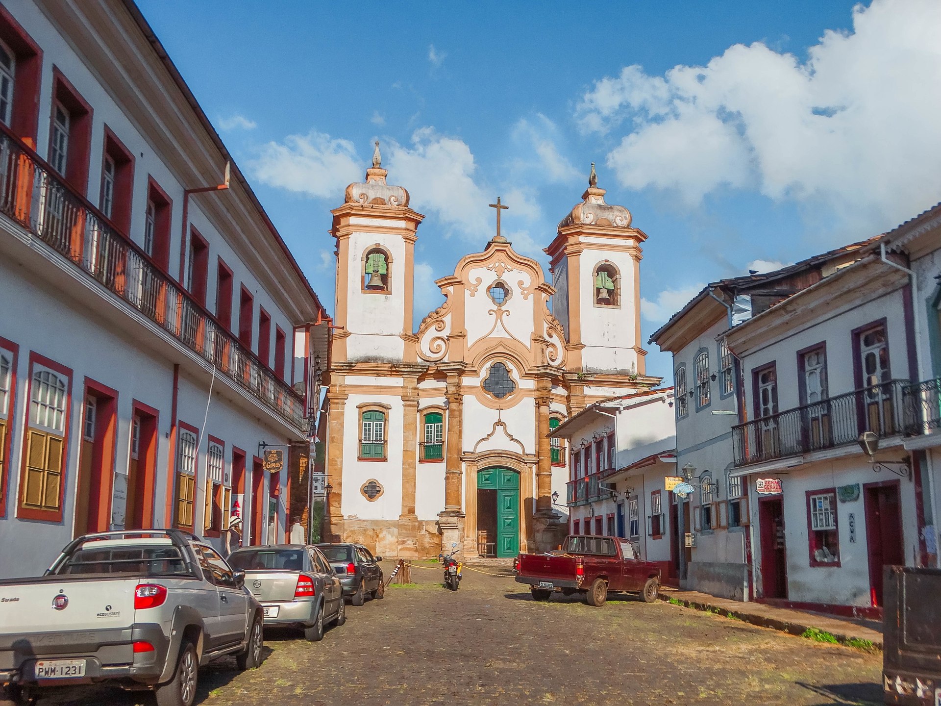 Vista da rua com a fachada da Igreja Matriz de Nossa Senhora do Pilar, em Ouro Preto. A imagem destaca a imponente estrutura barroca da igreja, com sua fachada ornamentada e detalhes arquitetônicos, situada no centro histórico da cidade
