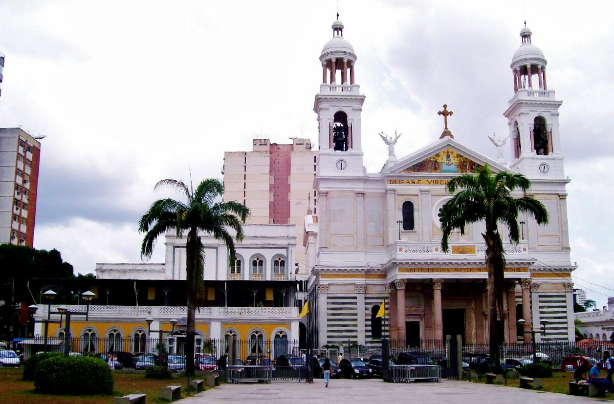Fachada frontal da Basílica Santuário de Nossa Senhora de Nazaré, em Belém (PA): edifício monumental de estilo neoclássico e eclético, com duas torres laterais que se elevam, frontão triangular, inscrições em latim, mosaicos regionais que retratam a Virgem como Rainha da Amazônia e colunas que emolduram uma entrada de aspecto monumental