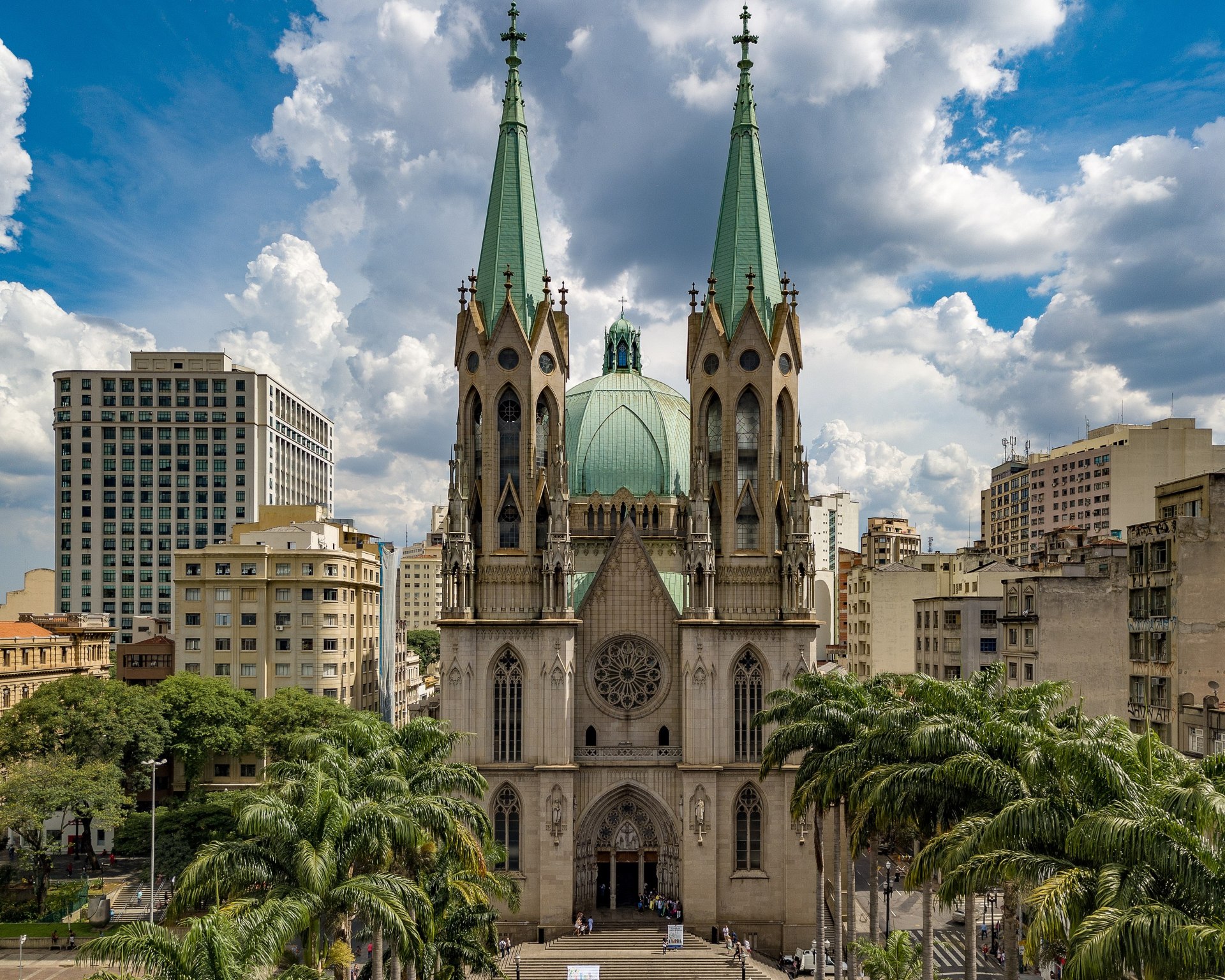 Vista frontal da Catedral Metropolitana de São Paulo, também conhecida como Catedral da Sé. A imagem destaca a fachada neogótica com rosácea central, duas torres imponentes e cúpula octagonal, rodeada por palmeiras imperiais e edificações urbanas. O céu claro e a luz suave ressaltam os detalhes arquitetônicos da igreja