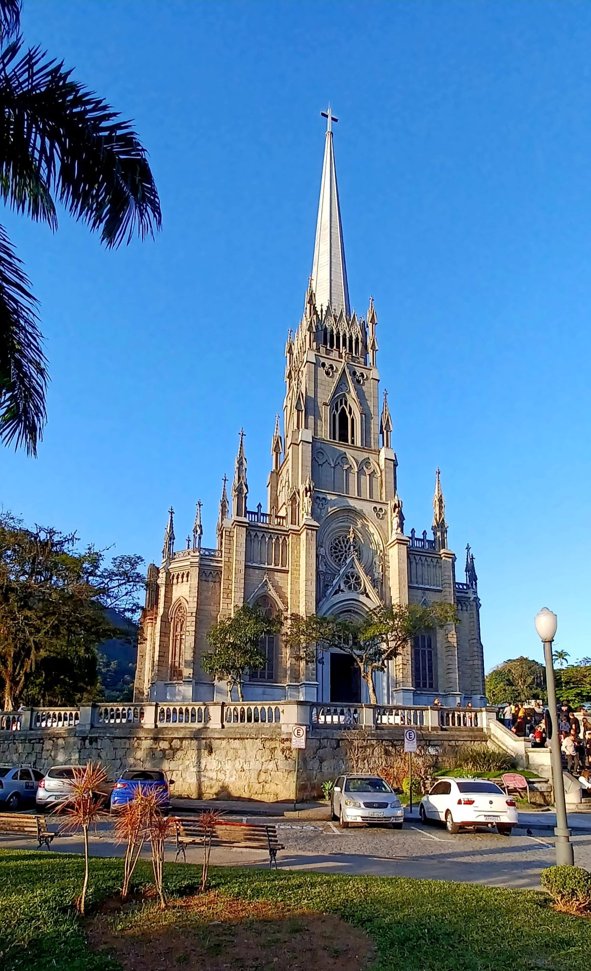Vista frontal da Catedral de São Pedro de Alcântara, em Petrópolis (RJ). A imagem destaca a fachada neogótica com rosácea central, portas de madeira ornamentadas e torres laterais. O entorno urbano e a vegetação ao redor complementam a composição arquitetônica