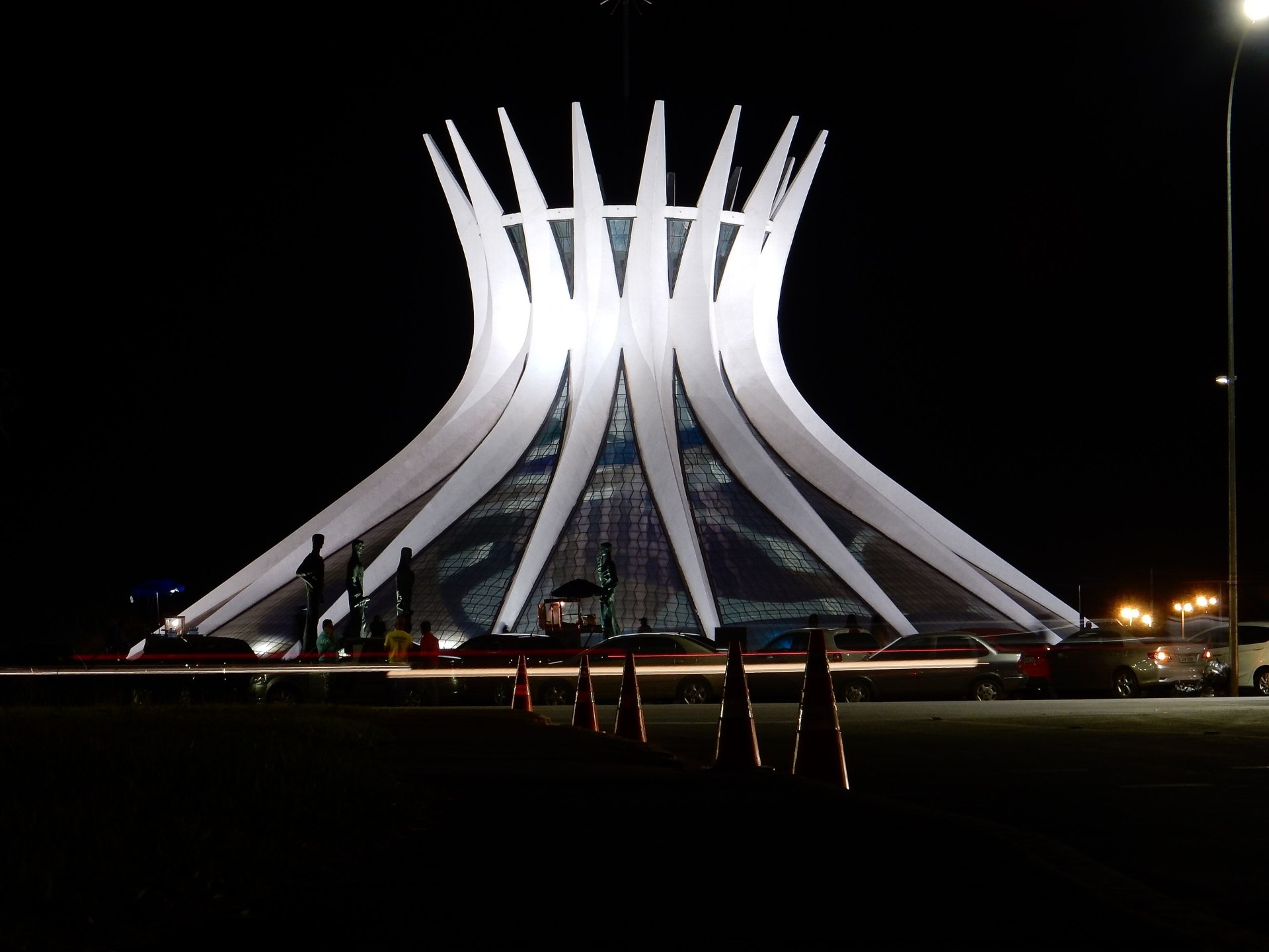 Vista externa da Catedral Metropolitana Nossa Senhora Aparecida, em Brasília (DF): estrutura modernista composta por 16 colunas de concreto que se erguem em forma de mãos unidas, com telhado cônico e vitrais coloridos. O edifício é rodeado por uma piscina refletora e está situado em uma praça aberta