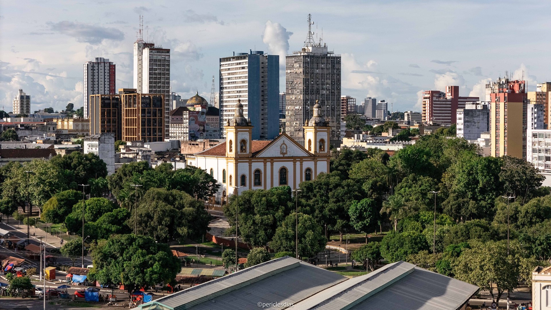 Fachada frontal da Catedral Metropolitana Nossa Senhora da Conceição, em Manaus (AM): edifício neoclássico em dois pavimentos, com três vãos centrais, frontão triangular, nicho com imagem sacra e torres laterais; fachadas em branco e detalhes em amarelo-ocre; escadaria frontal e área arborizada ao redor