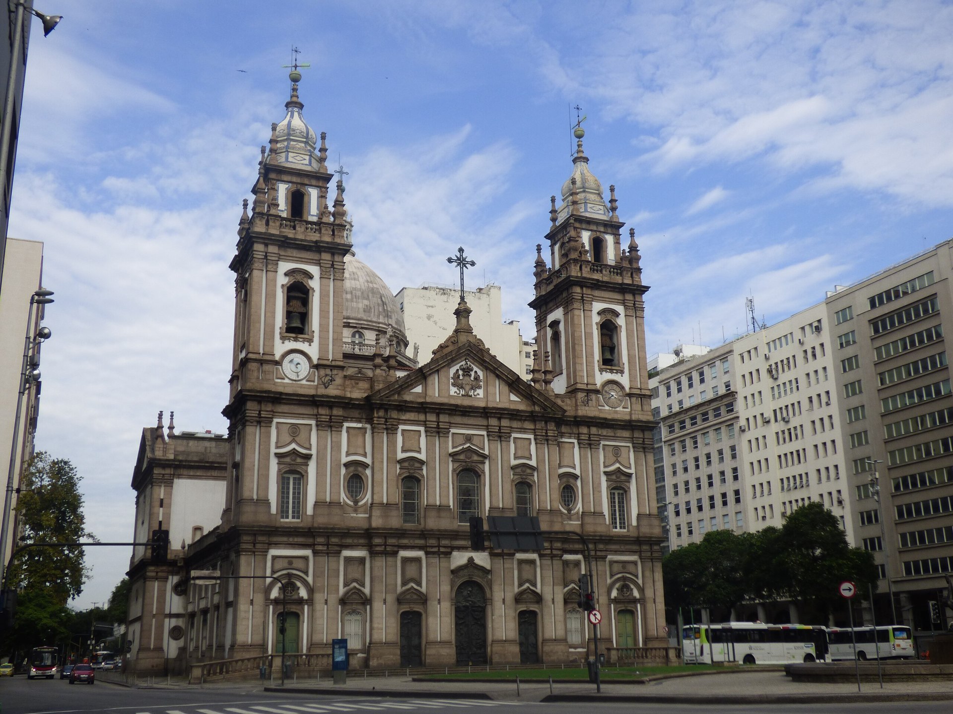 Vista frontal da Igreja de Nossa Senhora da Candelária, localizada na Praça Pio X, no Rio de Janeiro. A fachada neoclássica apresenta duas torres sineiras com relógios, portas de bronze esculpidas por Teixeira Lopes e detalhes arquitetônicos simétricos. O edifício é cercado por calçadas e vegetação, destacando-se no centro histórico da cidade
