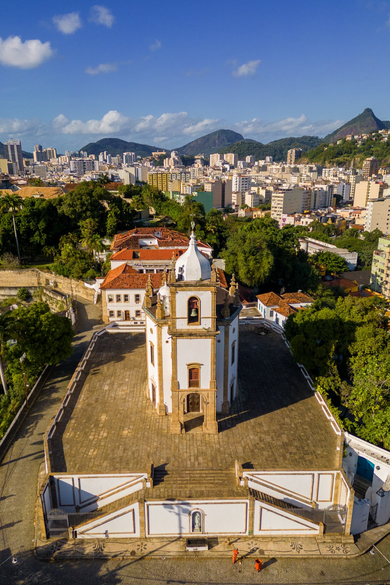 Vista aérea da Igreja de Nossa Senhora da Glória do Outeiro, situada no alto do Outeiro da Glória, no Rio de Janeiro. A imagem mostra a planta octogonal dupla da igreja, com a torre central e cúpula bulbosa, cercada por vegetação e edificações urbanas. O céu claro e a luz suave destacam os detalhes arquitetônicos da fachada e do entorno