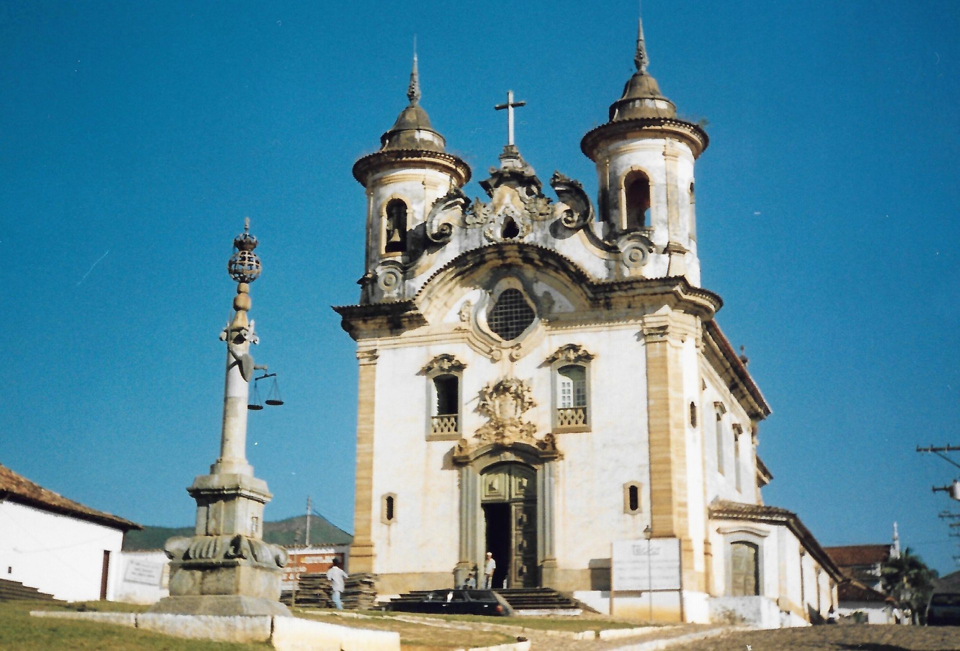 Vista frontal da Igreja de Nossa Senhora do Carmo, em Mariana (MG), destacando sua fachada rococó com torres cilíndricas recuadas e portada em pedra-sabão. O interior apresenta altar-mor com talha dourada rococó, projetado por Félix Antônio Lisboa. A igreja, restaurada após incêndio em 1999, é um ícone da arquitetura colonial brasileira