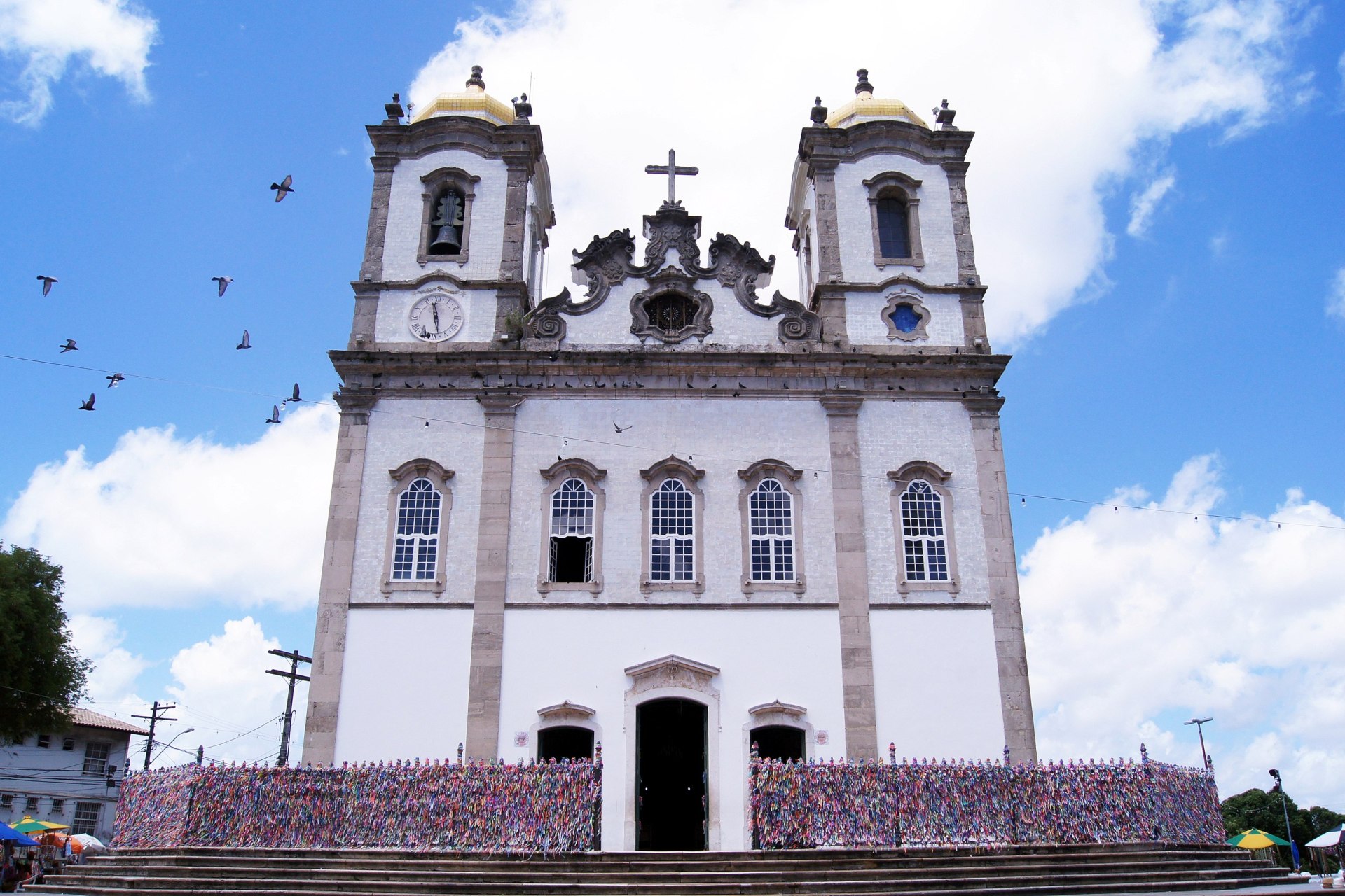 Fachada frontal da Igreja do Senhor do Bonfim, Salvador (BA): templo colonial em estilo neoclássico com fachada rococó parcialmente revestida de azulejos portugueses, duas torres sineiras laterais com cúpulas bulbosas, construída sobre uma colina, com escadaria ampla à frente