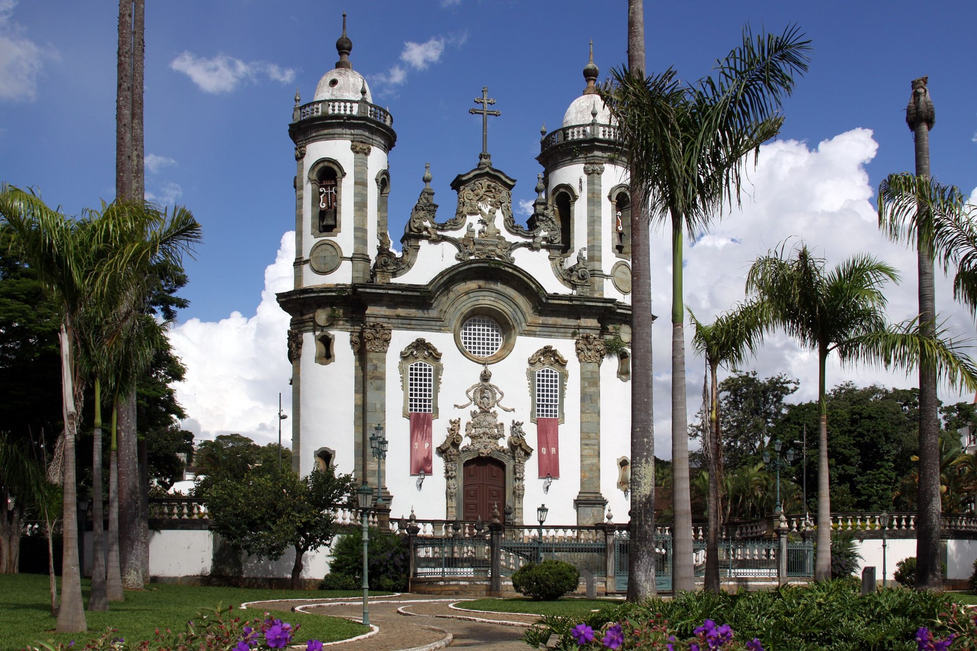 Vista frontal da Igreja de São Francisco de Assis, em São João del-Rei (MG), destacando sua fachada barroca com elementos rococós, incluindo a portada esculpida em pedra-sabão e o relevo de São Francisco adorando o Cristo Seráfico. O templo é cercado por um amplo adro com balaustrada de mármore branco e palmeiras imperiais, criando um ambiente de grande beleza e serenidade