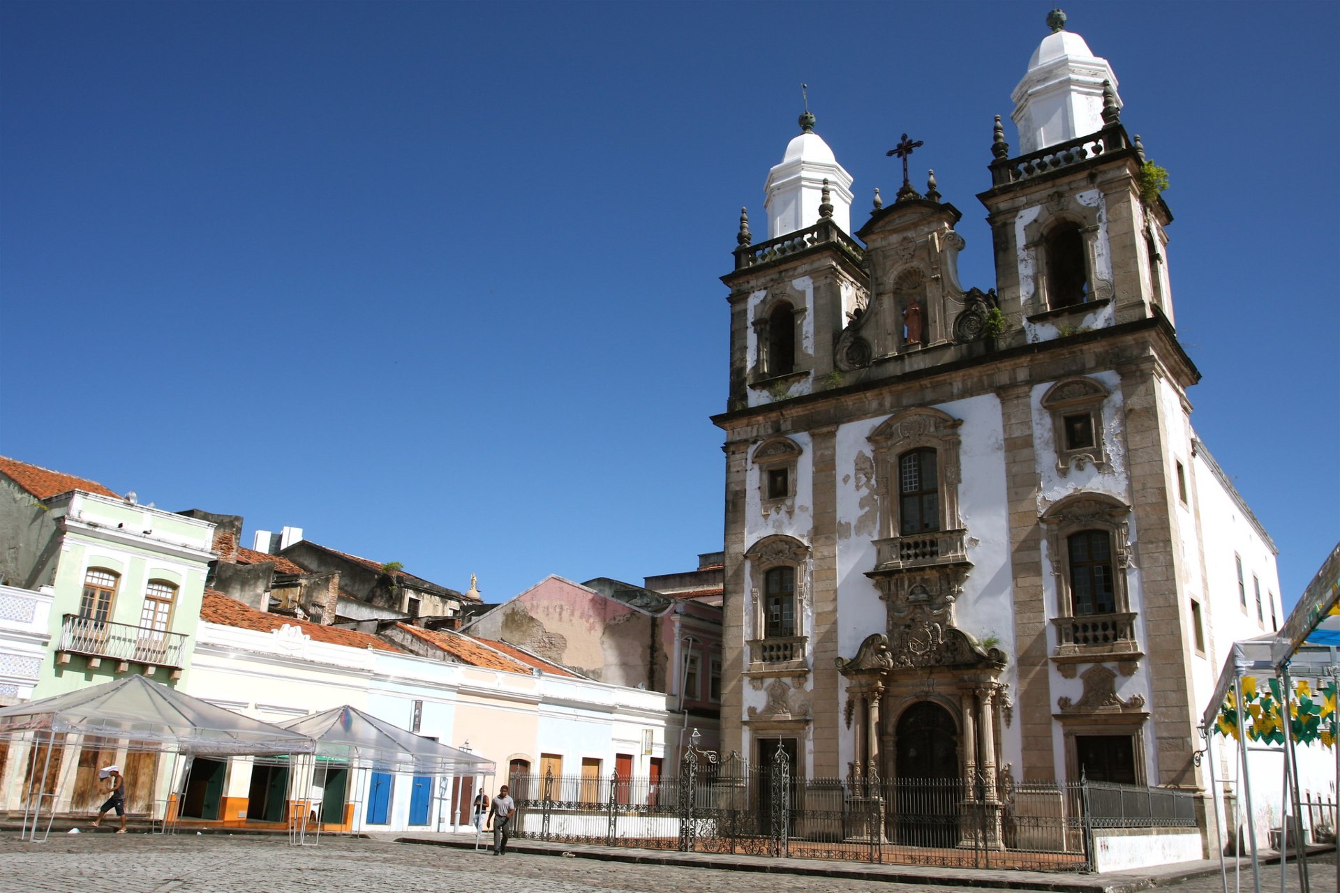 Fachada frontal da Igreja de São Pedro dos Clérigos, no Recife (PE): templo barroco de arenito, com duas torres laterais com pináculos, nicho central com imagem de São Pedro, elementos decorativos como volutas e sereias, e pátio em primeiro plano com casas coloniais ao redor
