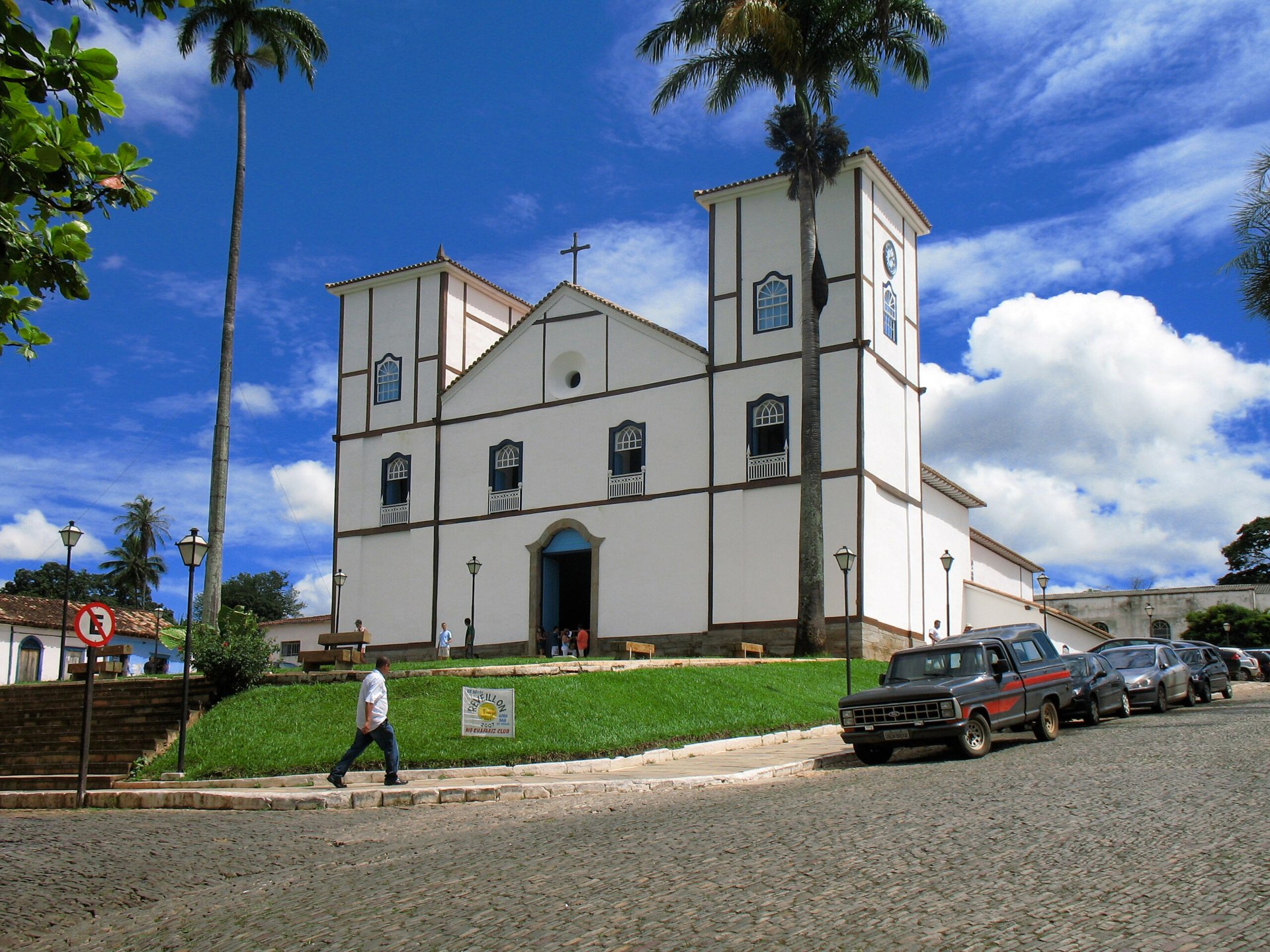 Vista frontal da Igreja Matriz de Nossa Senhora do Rosário, em Pirenópolis (GO): edifício colonial com fachada branca, torres sineiras e cruz central. O entorno é composto por escadarias e vegetação, destacando-se no centro histórico da cidade