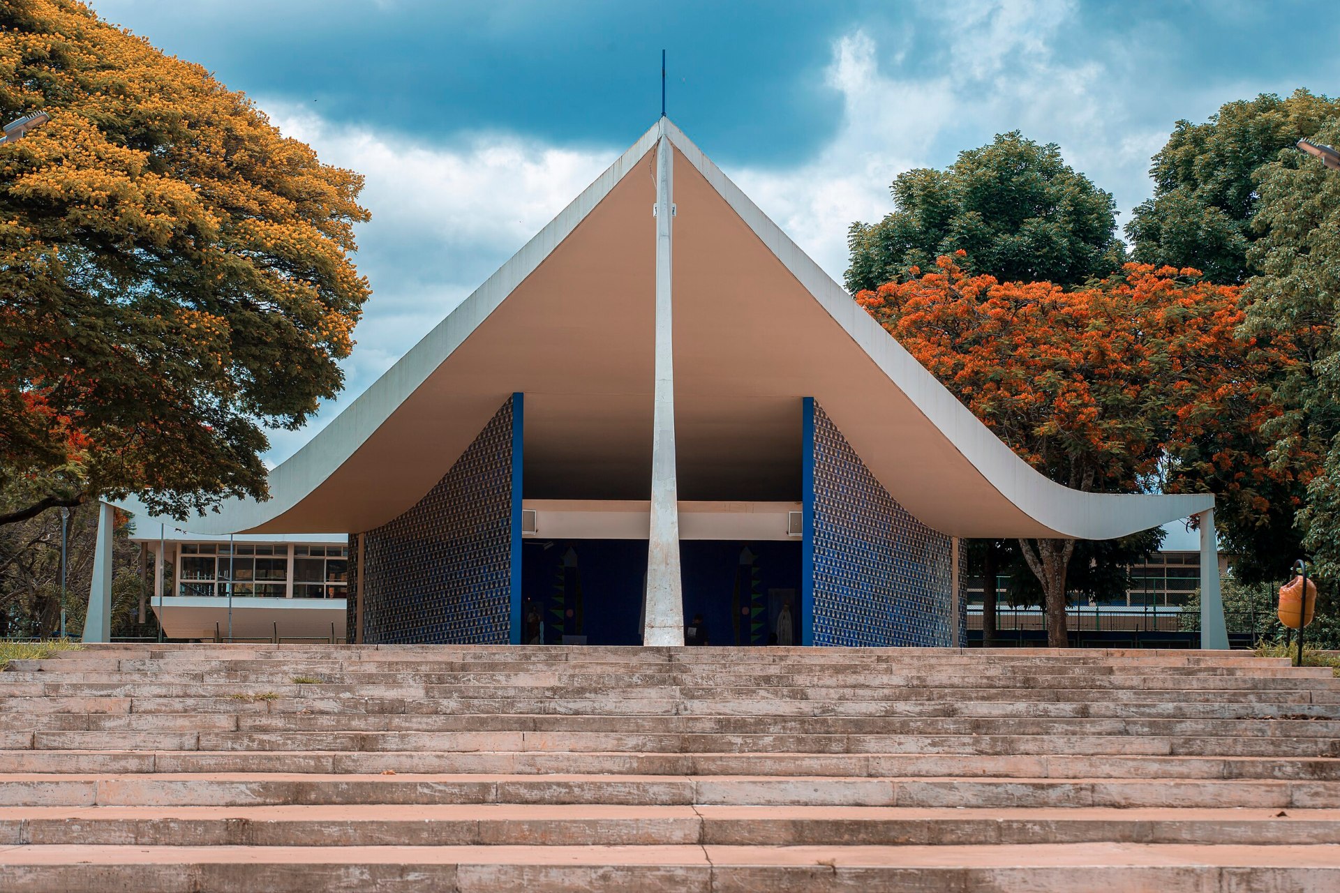 Vista frontal da Igreja Nossa Senhora de Fátima (Igrejinha), em Brasília (DF): estrutura modernista com telhado inclinado em forma de vela, paredes de concreto aparente e cruz central. A igreja está situada em uma praça ampla, cercada por vegetação