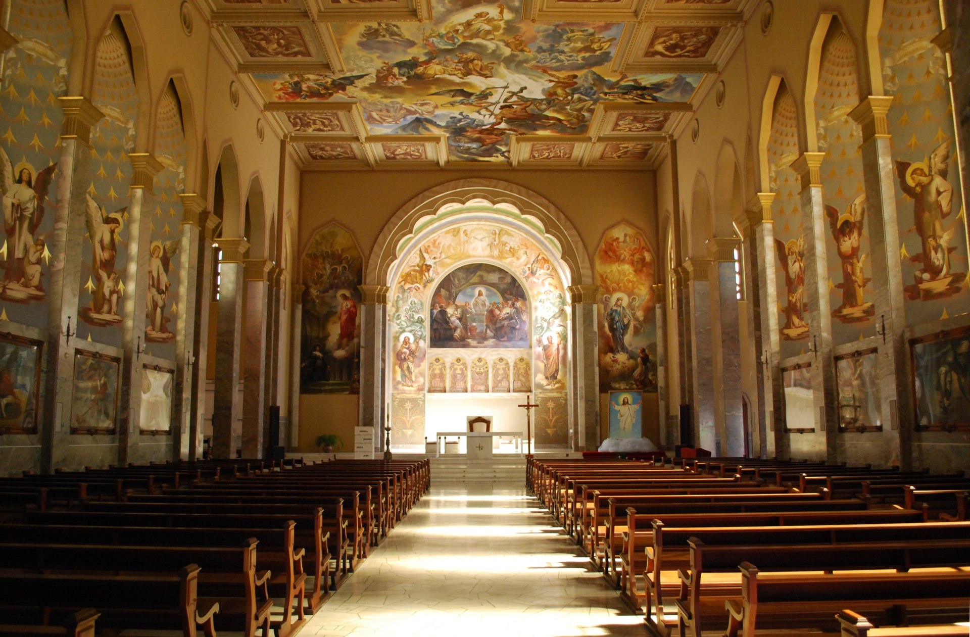 Interior da Igreja São Pelegrino mostra a ampla nave central com bancos de madeira alinhados, teto abobadado e detalhes arquitetônicos neogóticos. O altar é iluminado por vitrais coloridos que projetam luz natural no ambiente, realçando a riqueza dos ornamentos e a atmosfera de reverência e tranquilidade do espaço religioso