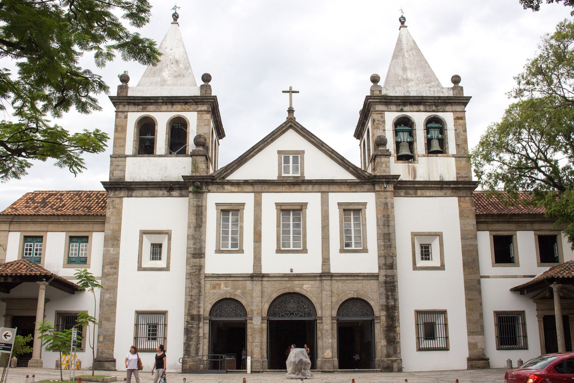 Vista frontal do Mosteiro de São Bento, no Rio de Janeiro: fachada austera em estilo maneirista com três arcos de entrada e frontão triangular. Flanqueada por duas torres coroadas por pináculos piramidais, a entrada é ladeada por portões de ferro do século 19. O edifício está situado no alto do Morro de São Bento, cercado por vegetação e edificações urbanas