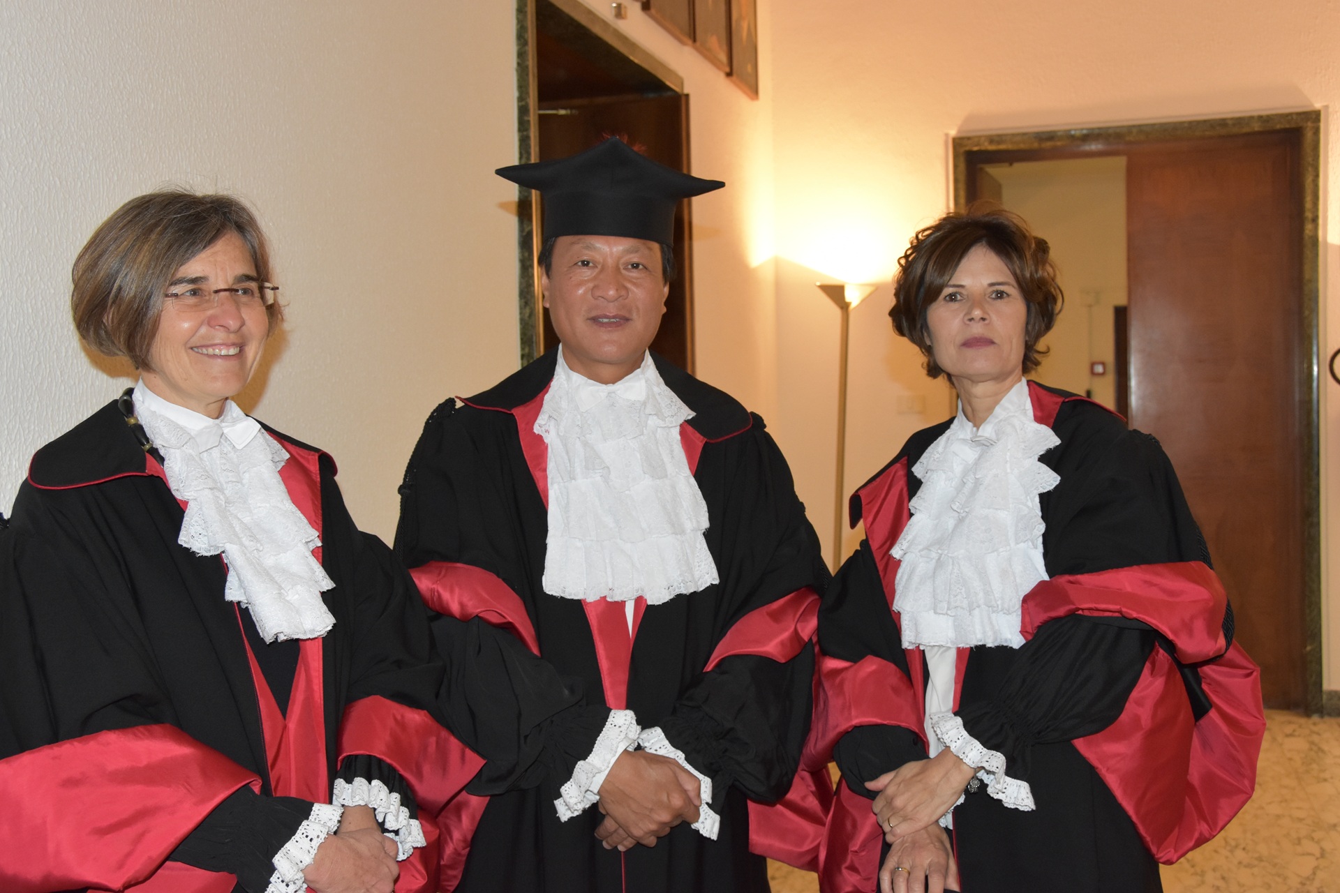 Três pessoas vestindo becas acadêmicas com detalhes em vermelho e branco, posando em um corredor iluminado. No centro, está Kongjian Yu, durante a cerimônia em que recebeu o título de Doutor Honoris Causa na Universidade Sapienza de Roma, em 2017