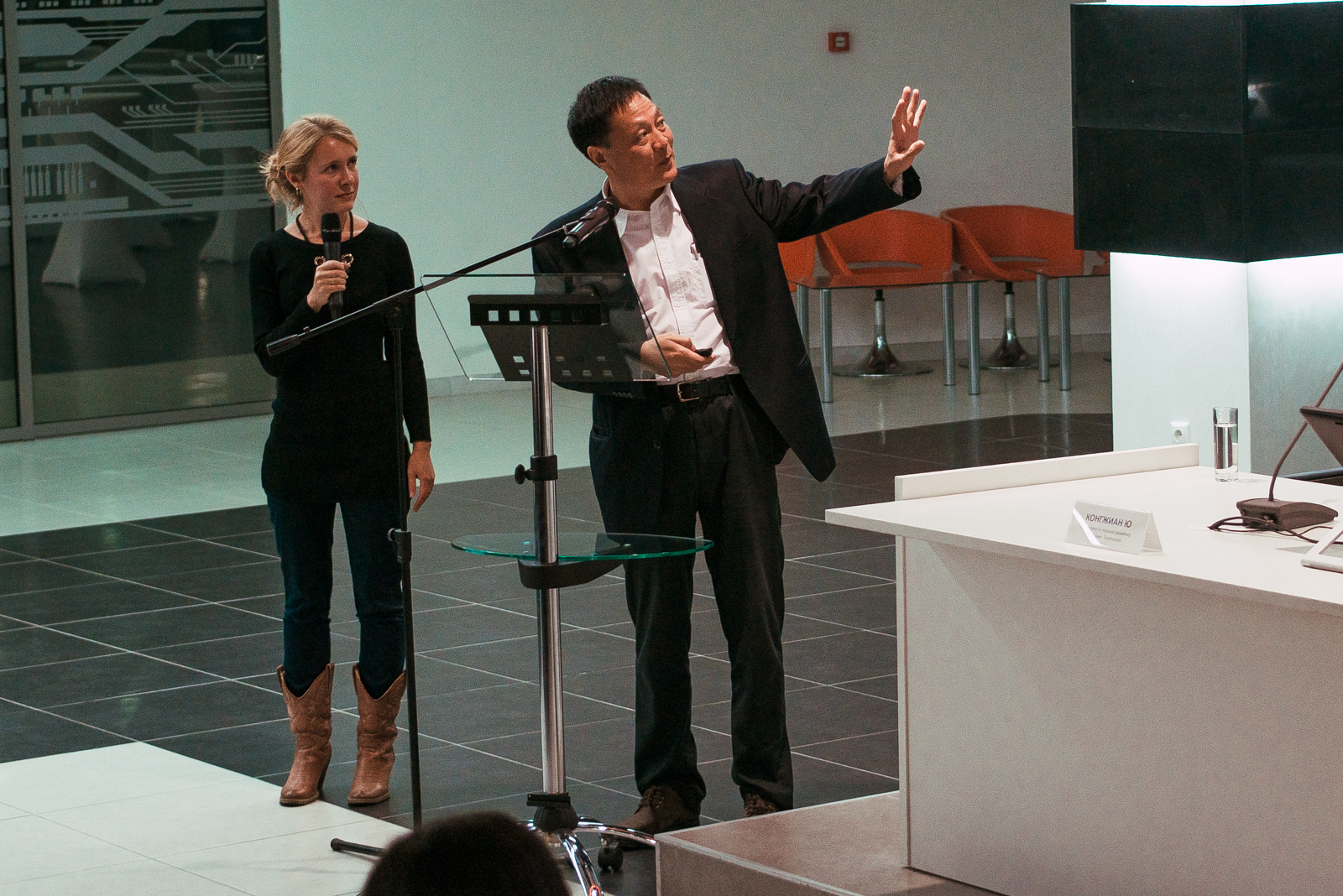 Kongjian Yu aparece em pé, ao lado de uma mulher, durante uma palestra. Ele gesticula enquanto fala, com um microfone, em um auditório moderno. Na mesa à frente, uma placa com seu nome pode ser vista
