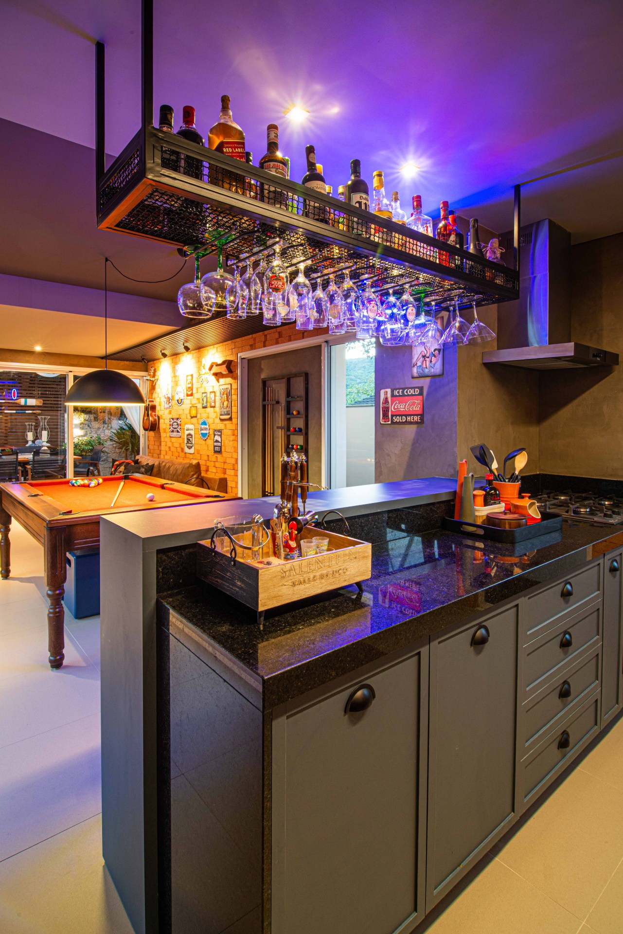 Balcão gourmet cinza com pedra preta. Acima há um prateleira suspensa com bebidas e taças. Ao fundo, uma mesa de bilhar