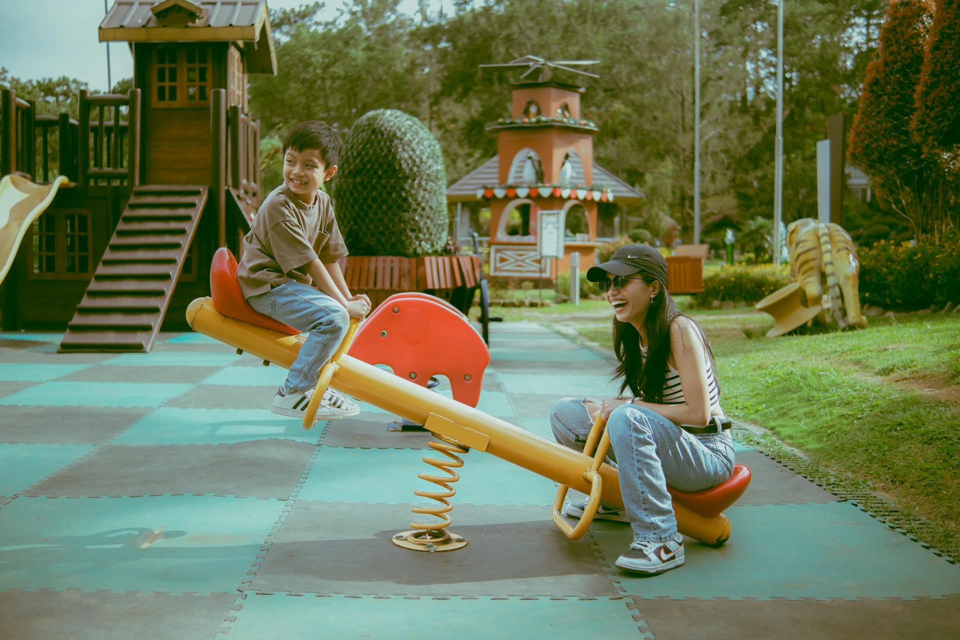 Criança e mulher se divertem em uma gangorra amarela em um parque infantil ao ar livre, com brinquedos de madeira e áreas verdes ao fundo