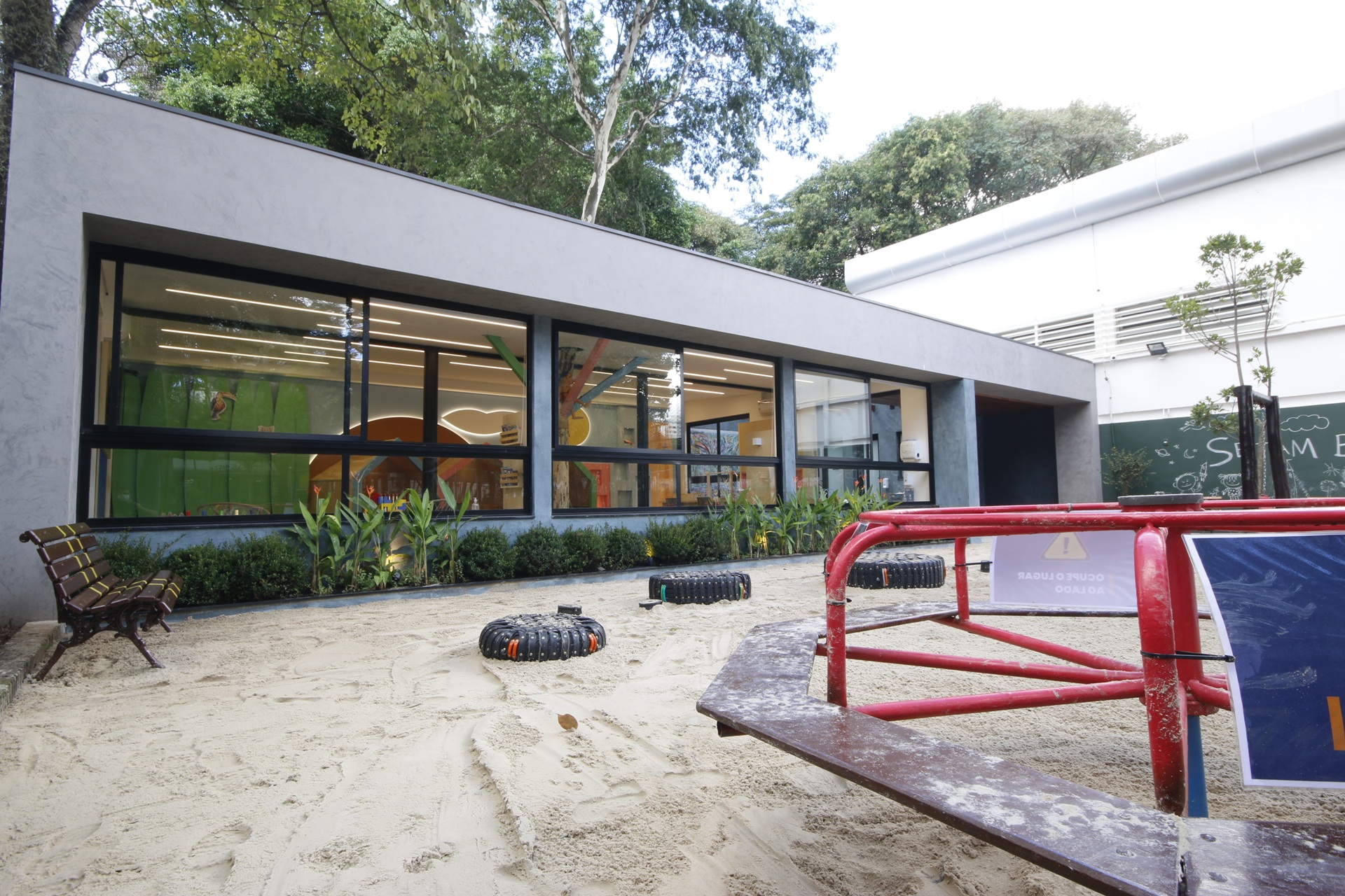 Parque infantil com área de areia, pneus reutilizados como brinquedos e estrutura de ferro vermelha em frente a um prédio moderno, destacando a relevância do planejamento arquitetônico no projeto de espaços infantis