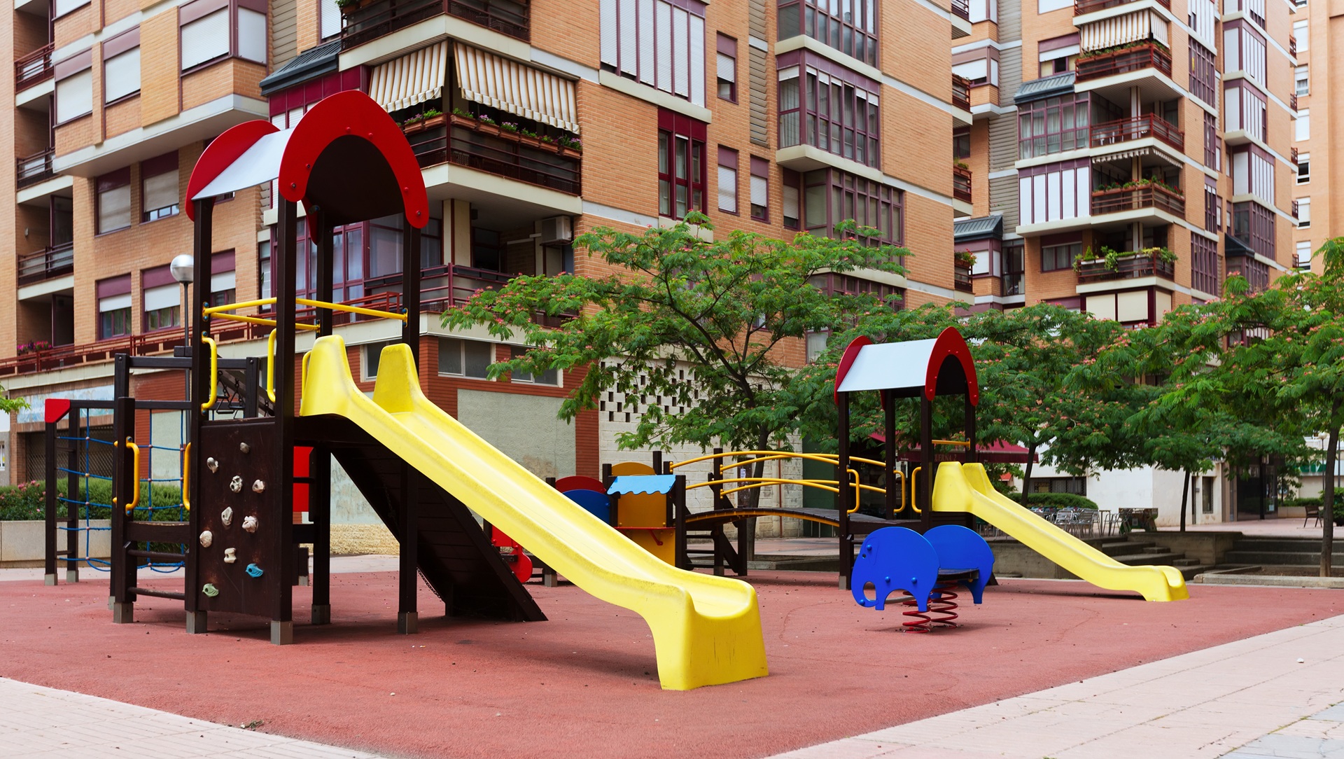 Playground em área urbana com piso emborrachado vermelho, escorregadores amarelos, brinquedo de mola em formato de elefante azul e árvores ao redor, em frente a prédios residenciais