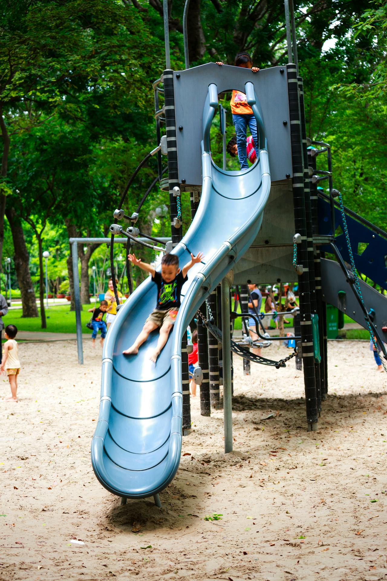 Criança desliza em escorregador curvo de metal em parque infantil ao ar livre com piso de areia, enquanto outras crianças brincam ao fundo