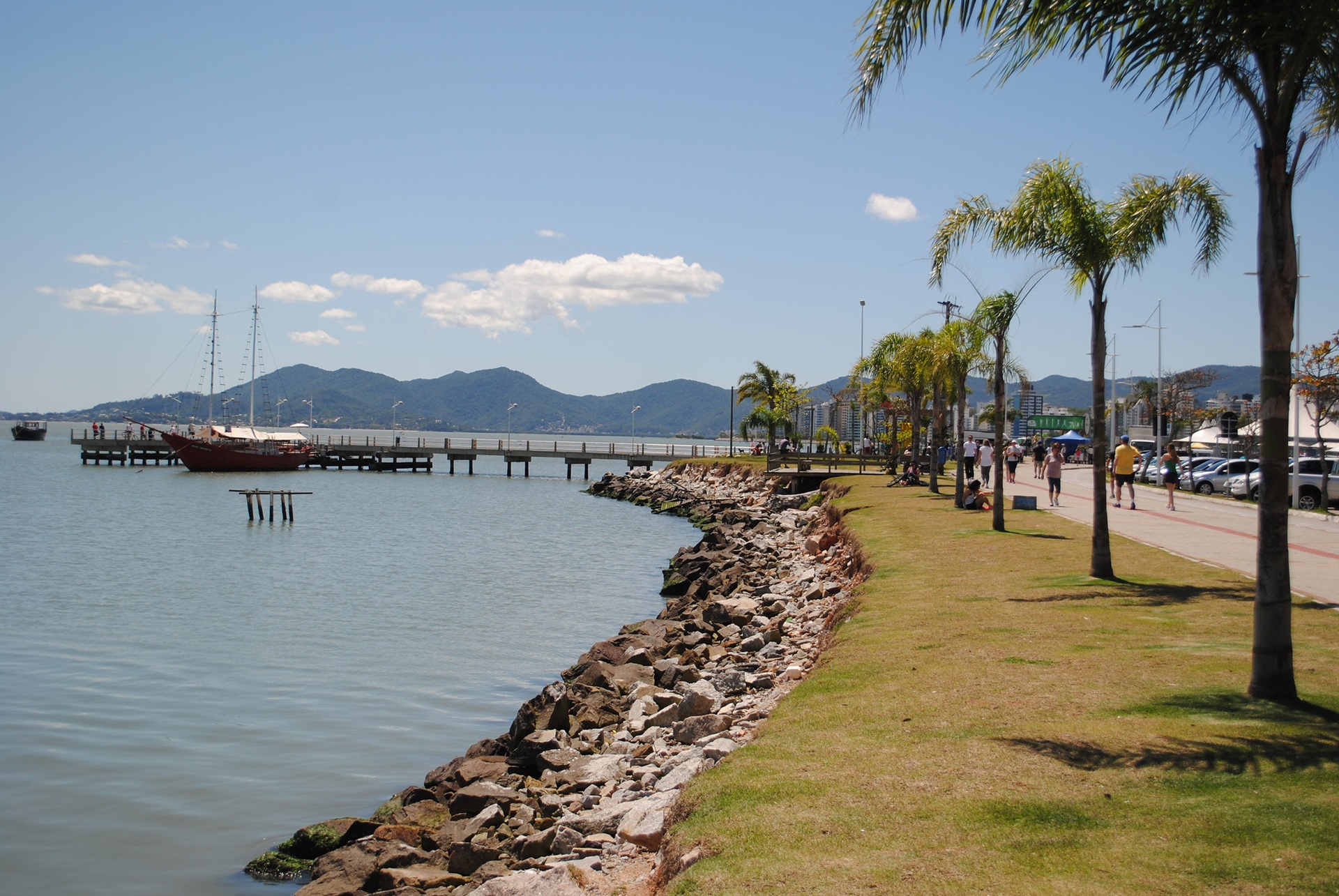 Vista da Avenida Beira-Mar Norte, em Florianópolis (SC), com calçadão arborizado, pista de caminhada, ciclovia, bancos e áreas de lazer à beira da Baía Norte; ao fundo, mar, embarcações, prédios e montanhas compõem a paisagem integrada entre urbanismo e natureza