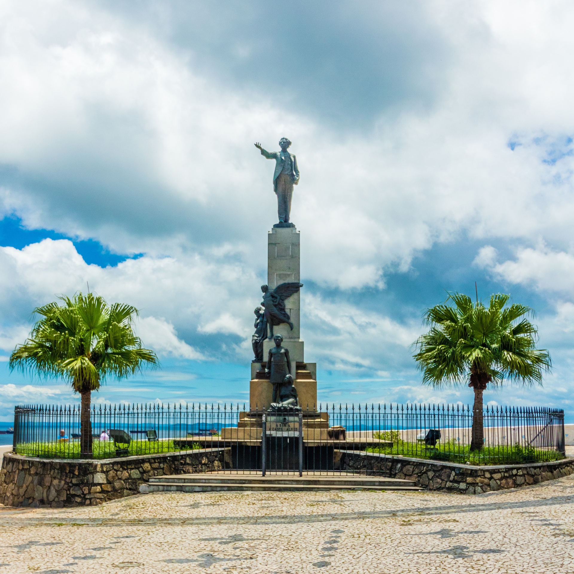 Monumento a Castro Alves na Praça Castro Alves, na Avenida Sete de Setembro, em Salvador (BA); a escultura mostra o poeta em posição altiva sobre pedestal ladeado por figuras simbólicas, cercado por gradil, palmeiras e calçamento em mosaico, com vista para a Baía de Todos-os-Santos