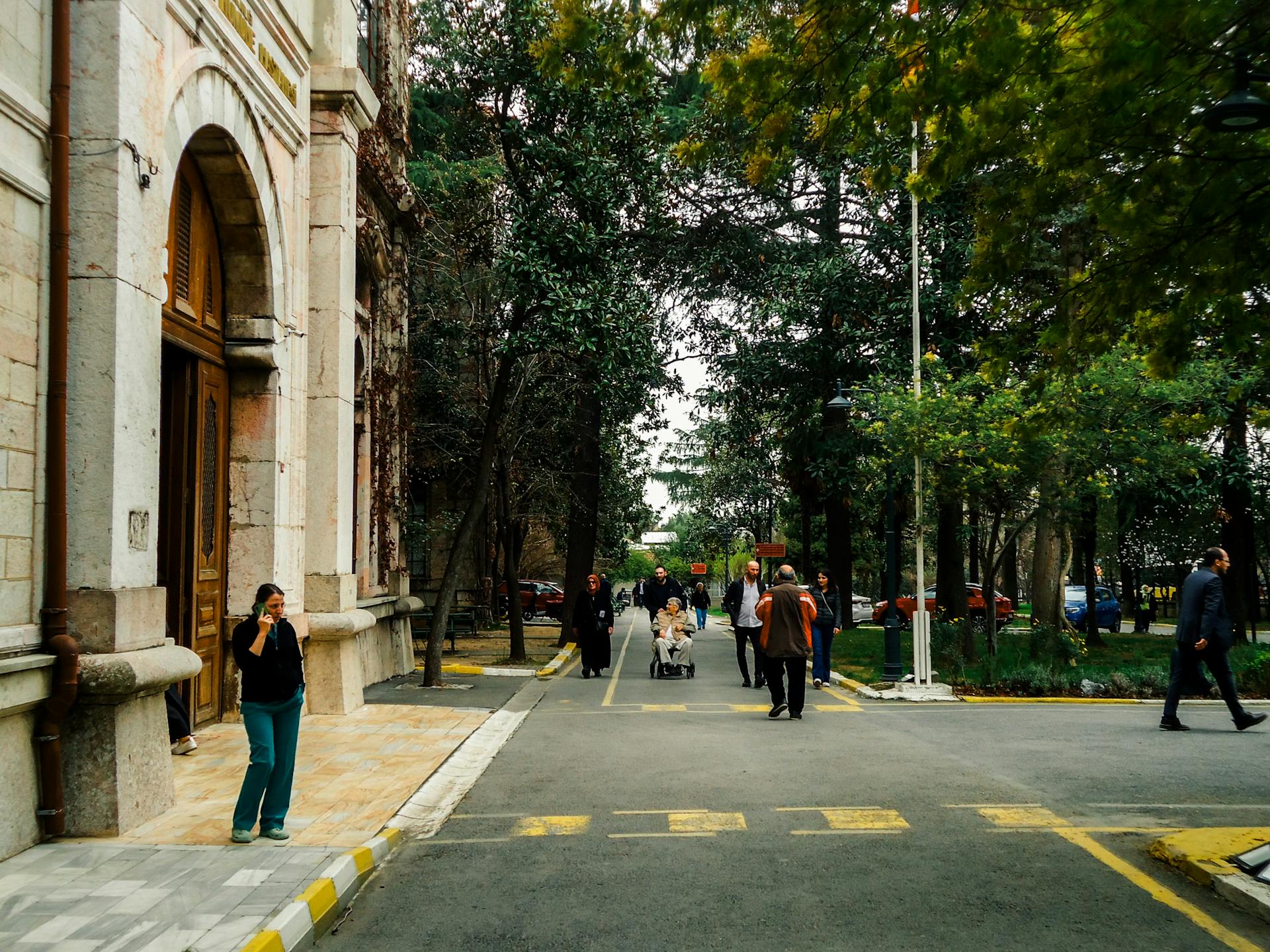 Pessoa caminha falando ao celular enquanto outras atravessam a rua em frente a um prédio histórico. O cenário urbano é cercado por árvores e movimento tranquilo de pedestres