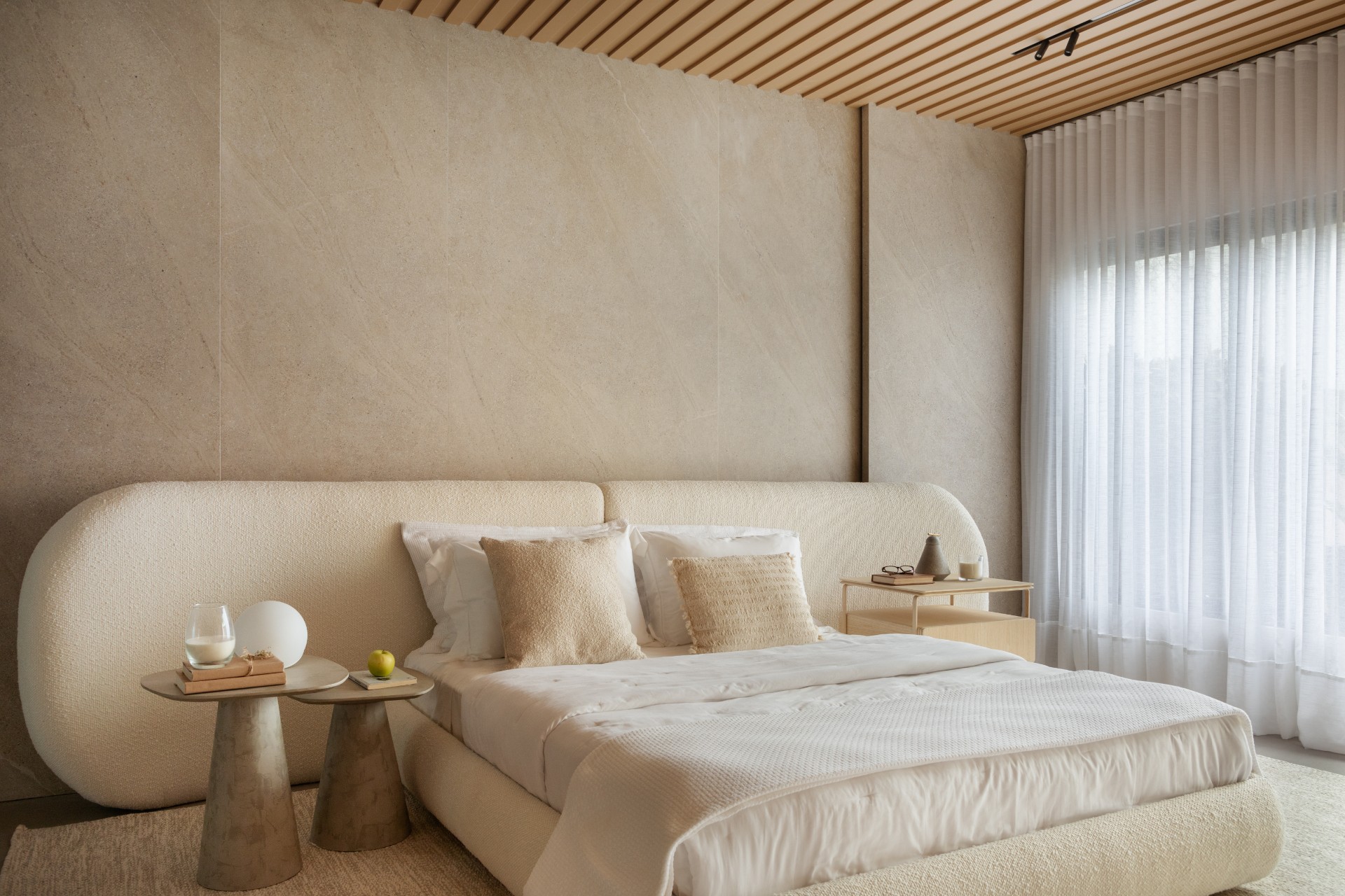 Quarto com cama de formas curvas em tecido bege, cabeceira ampla, paredes e piso em tons claros e cortina de linho