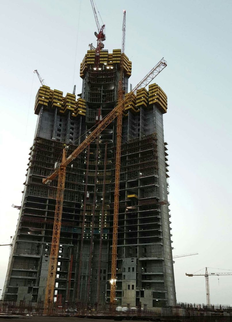 Imagem da Jeddah Tower em construção em 2016. A foto mostra a base larga e o núcleo central robusto da torre, rodeado por guindastes e andaimes. A estrutura em formato triangular foi projetada para oferecer máxima estabilidade e resistência aos ventos intensos em grandes altitudes
