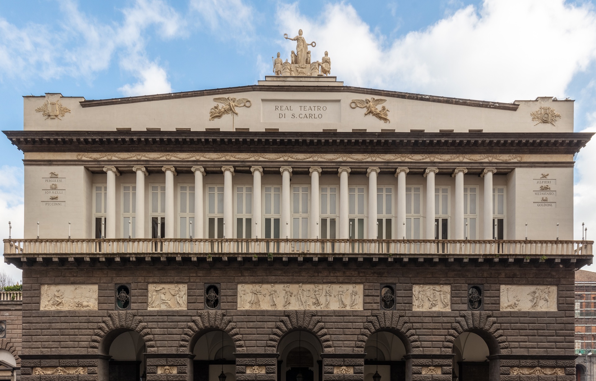 casa de opera Fachada do Teatro San Carlo, em Nápoles, Itália, com colunas brancas, esculturas no topo e relevos ornamentais na parte inferior, sob um céu azul com nuvens suaves.