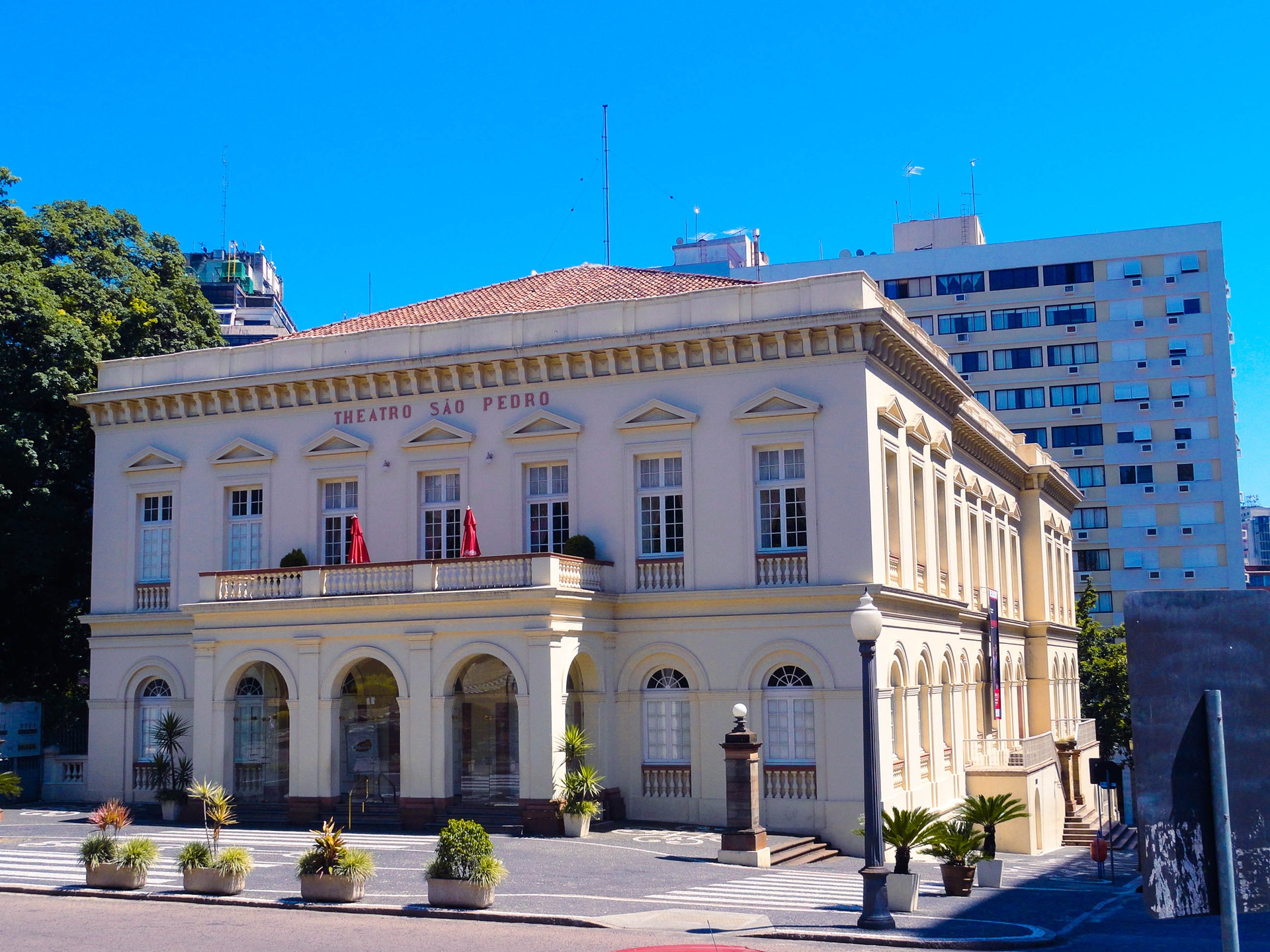 casa de ópera Fachada do Theatro São Pedro, em Porto Alegre, com arquitetura clássica em tons claros, janelas simétricas e arcos na entrada, rodeado por plantas em vasos e edifícios ao fundo, sob um céu azul intenso.