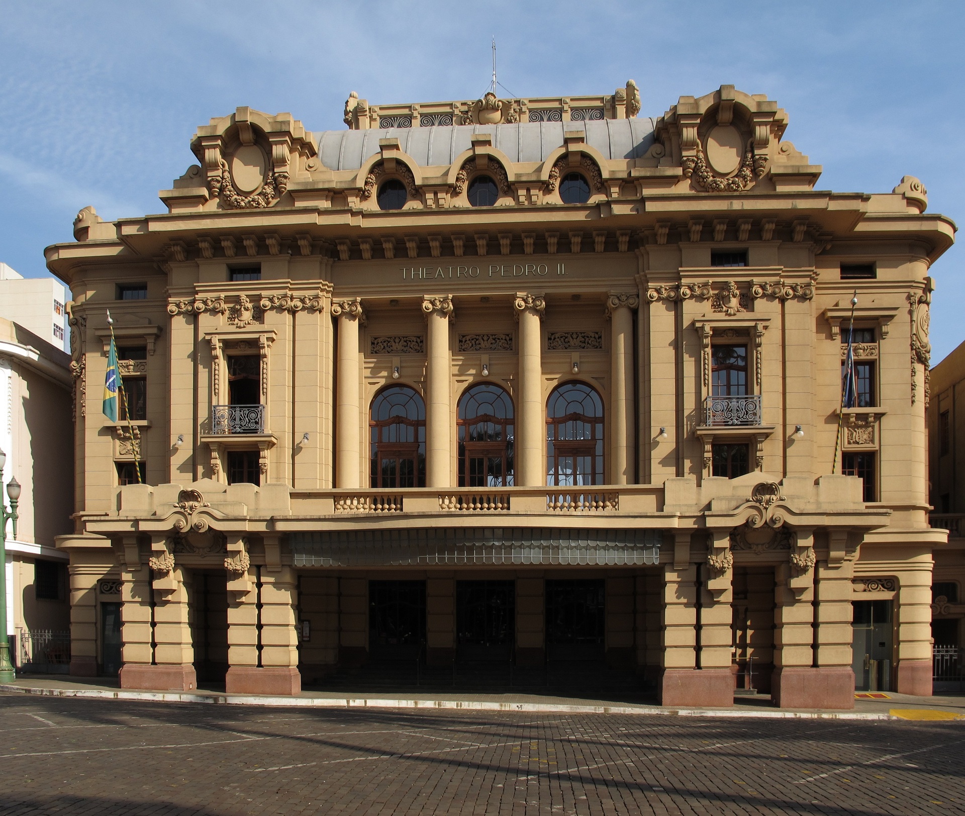 casa de ópera Fachada do Theatro Pedro II, em Ribeirão Preto, com arquitetura art déco em tons terrosos, colunas imponentes e bandeiras do Brasil e de São Paulo hasteadas nas laterais, sob céu claro.