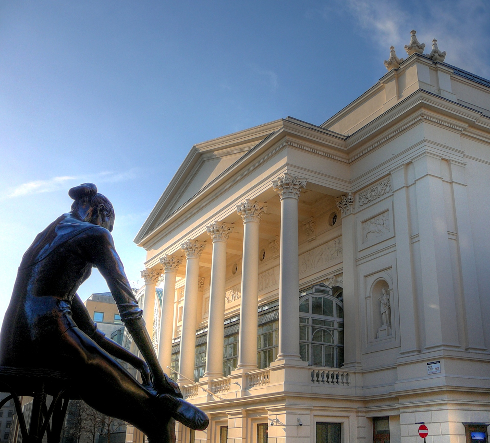 casa de ópera Fachada da Royal Opera House, em Londres, com colunas clássicas e esculturas ornamentais, vista ao entardecer. Em primeiro plano, uma estátua de bailarina sentada, criando conexão visual com o teatro.