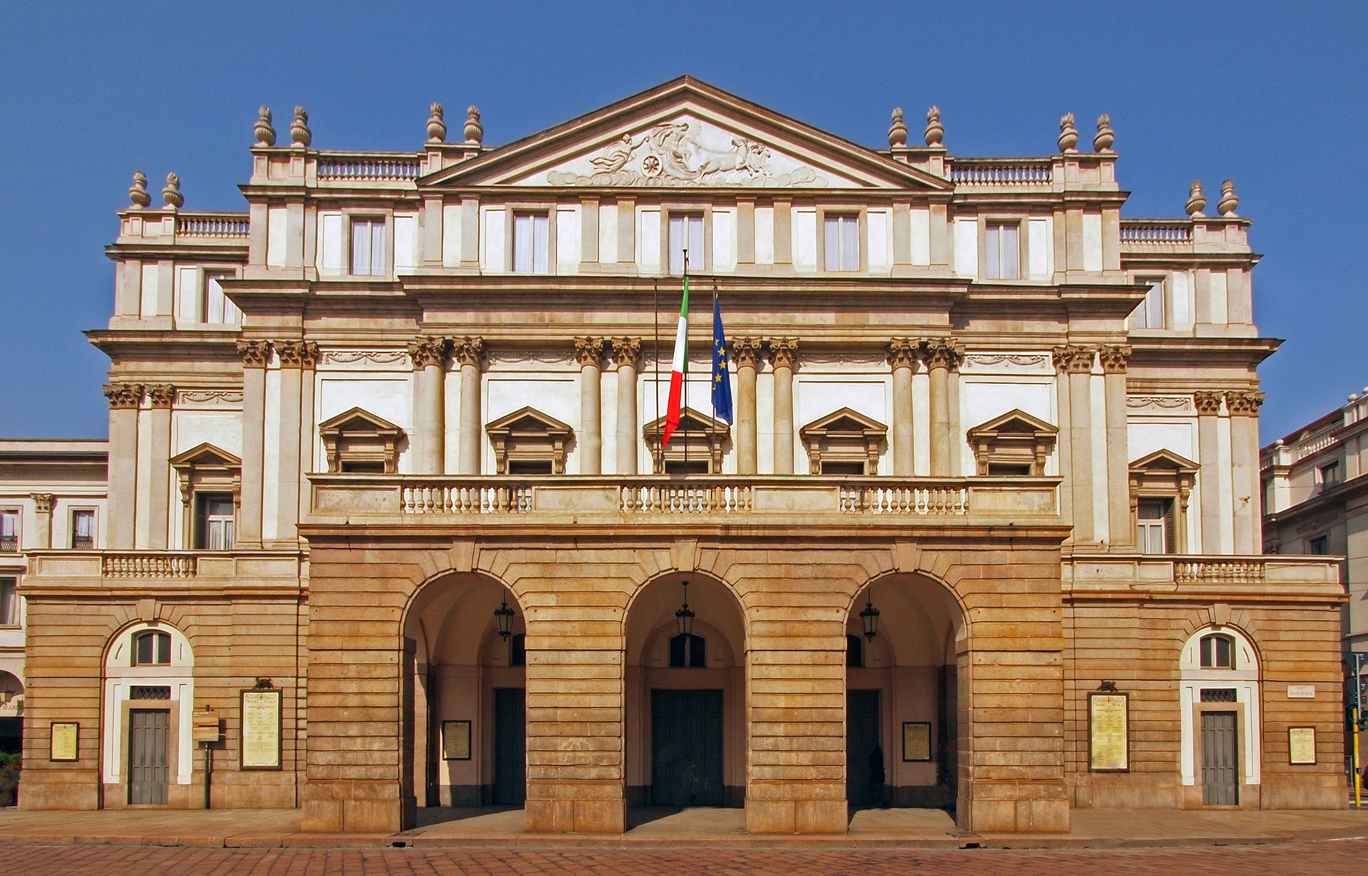 casa de ópera Fachada do Teatro alla Scala, em Milão, Itália, com arquitetura clássica, colunas imponentes, bandeiras da Itália e da União Europeia, sob um céu azul sem nuvens.