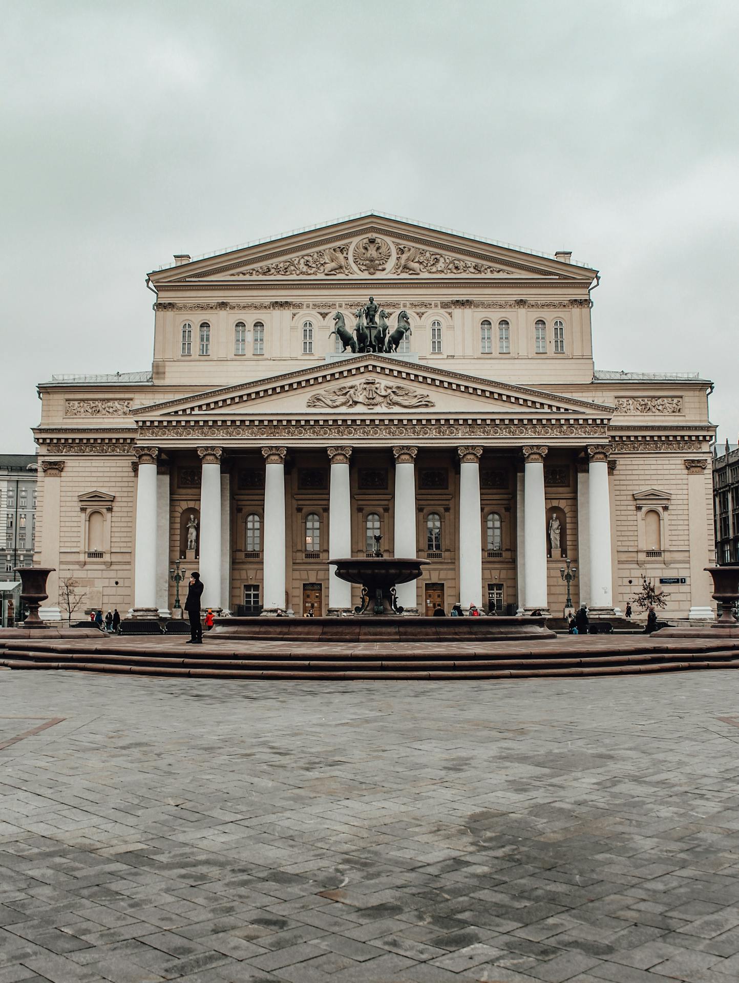 casa de ópera Fachada do Teatro Bolshoi, em Moscou, Rússia, com colunas brancas imponentes, esculturas em relevo e uma quadriga no topo, vista a partir de uma praça com fonte central em primeiro plano.