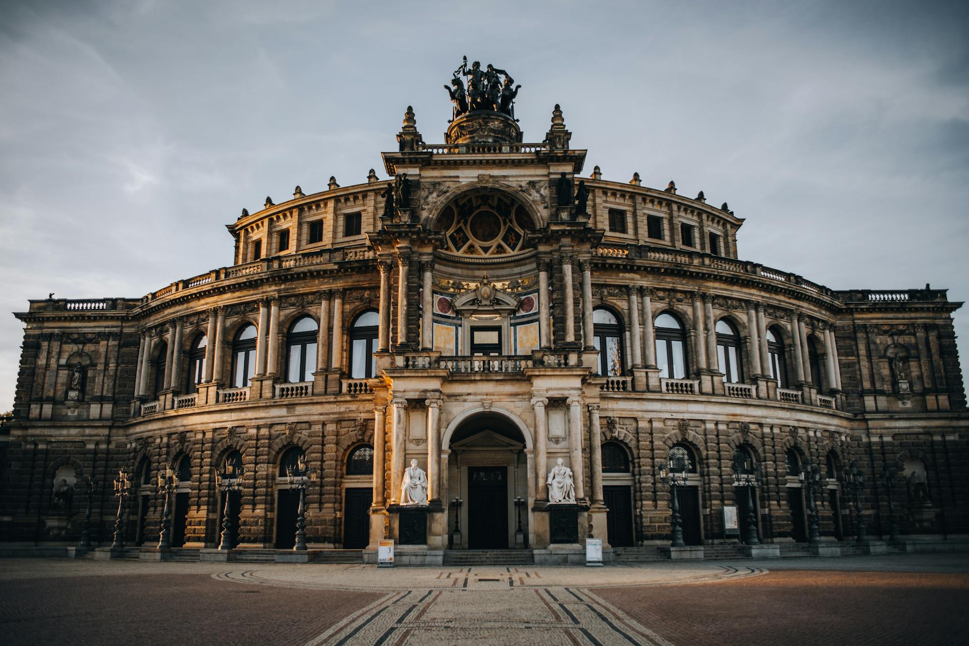 casa de ópera Fachada da Ópera Semper, em Dresden, Alemanha, com arquitetura circular imponente, esculturas no topo e duas estátuas brancas ao lado da entrada principal, iluminada pela luz suave do entardecer.