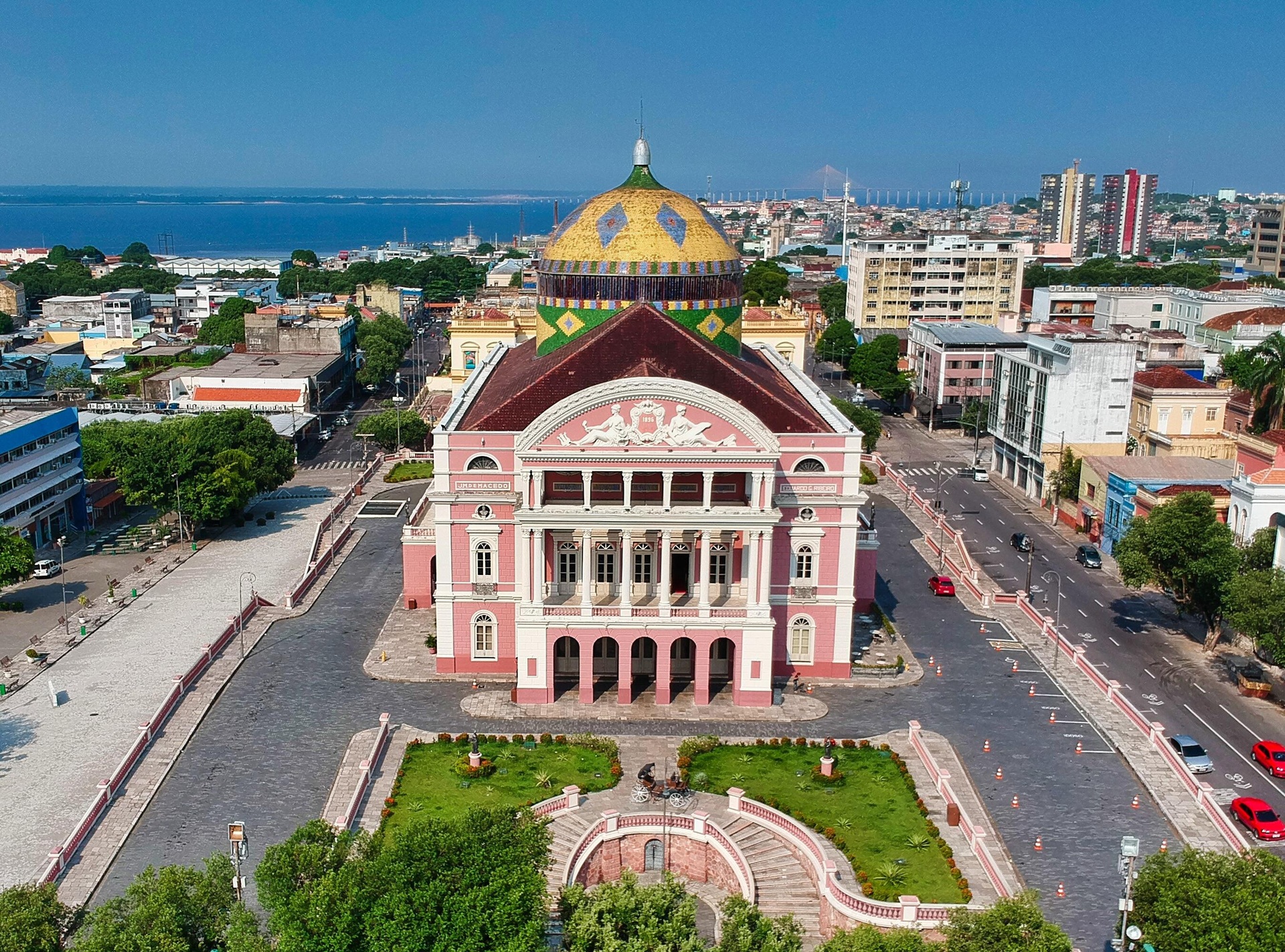 casa de ópera Vista aérea do Teatro Amazonas, em Manaus, com fachada rosa e cúpula colorida nas cores da bandeira do Brasil, cercado por ruas e prédios da cidade, com o Rio Negro visível ao fundo.