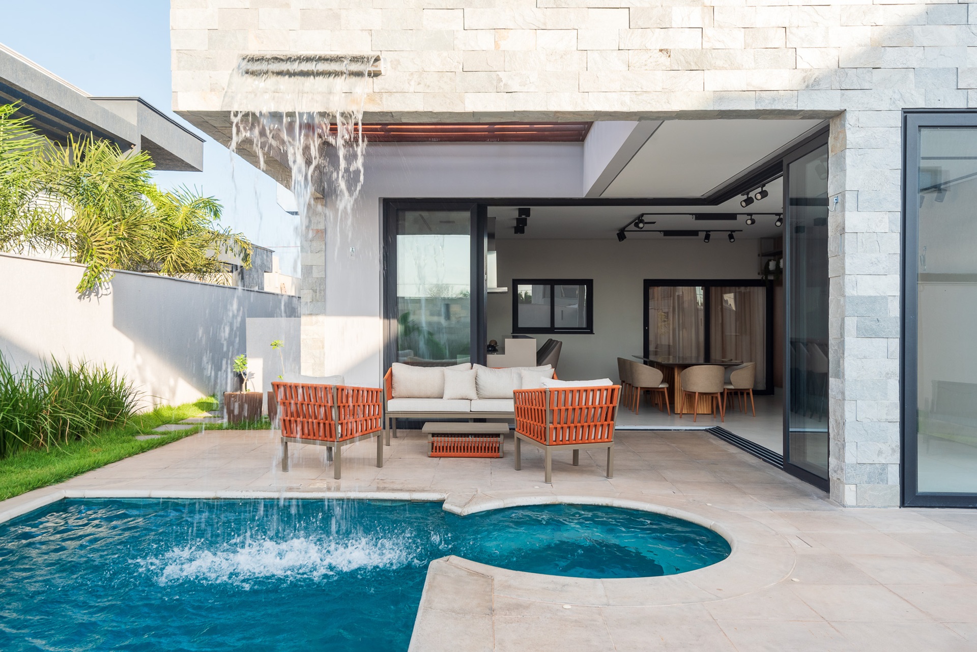 Área externa com revestimento em tons claros e piscina de alvenaria azul com cascata suspensa