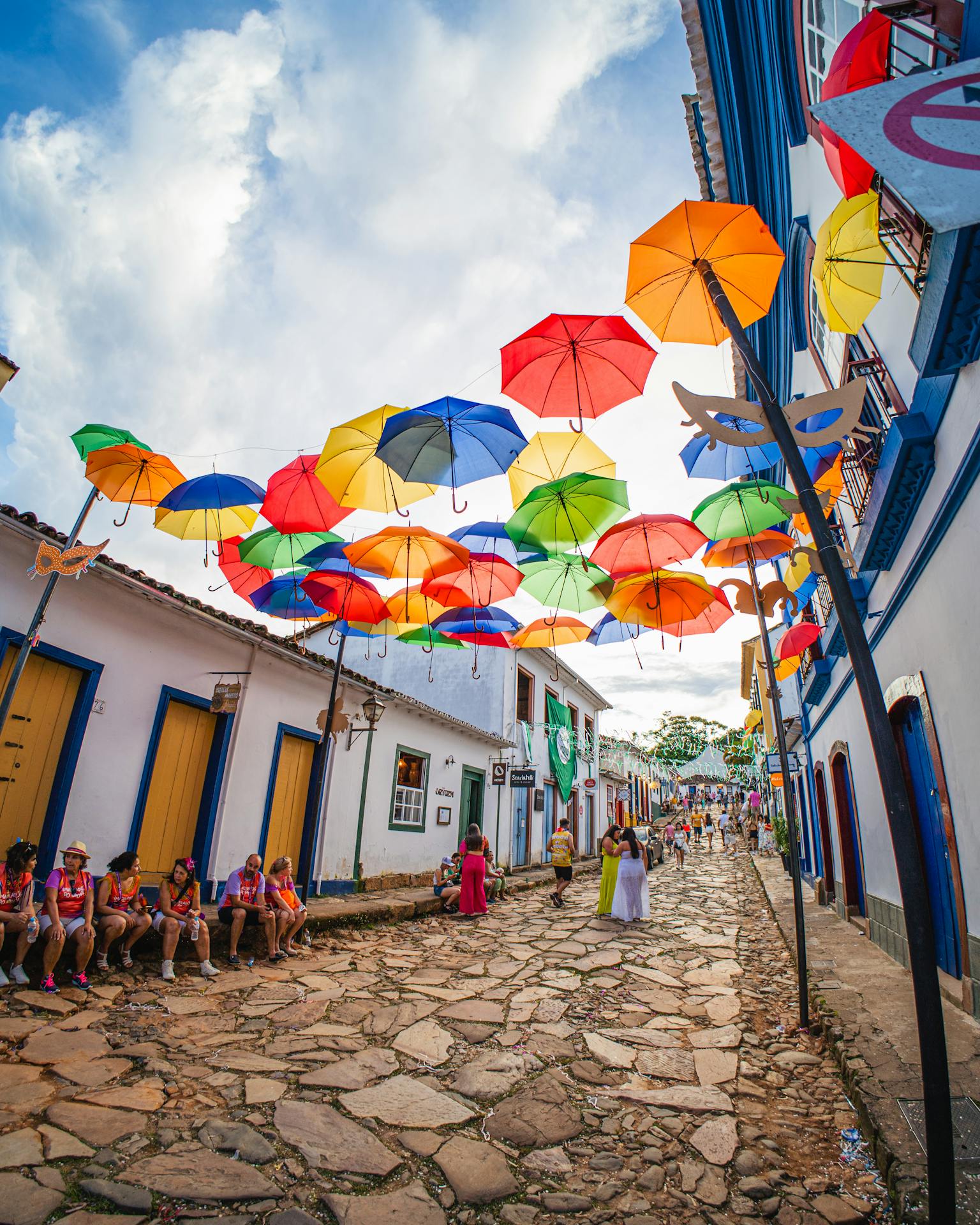 Rua histórica de Tiradentes, Minas Gerais, decorada com guarda-chuvas coloridos durante o Carnaval, com pessoas circulando e festejando
