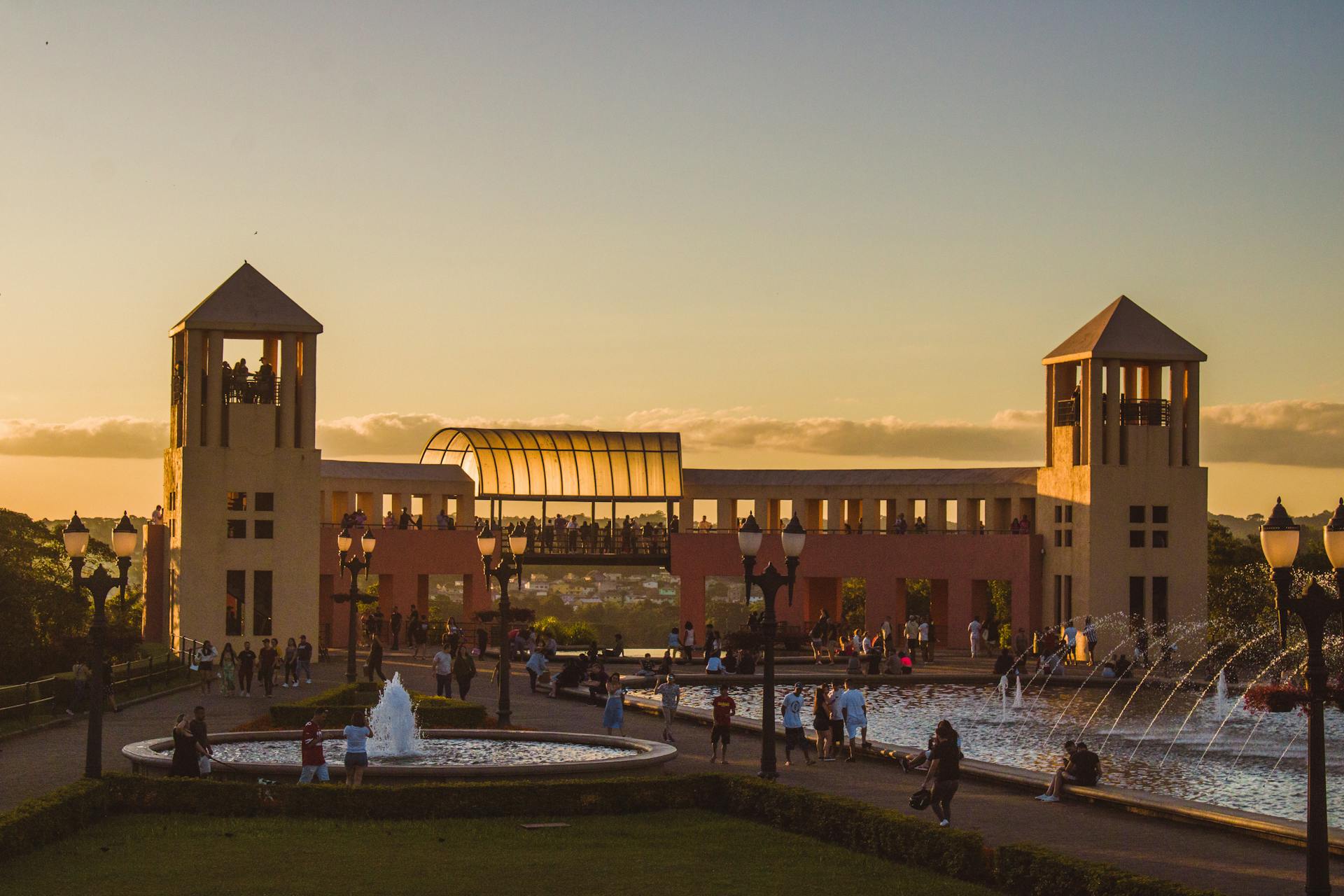 Parque Tanguá, em Curitiba, com pessoas caminhando e fontes iluminadas ao entardecer