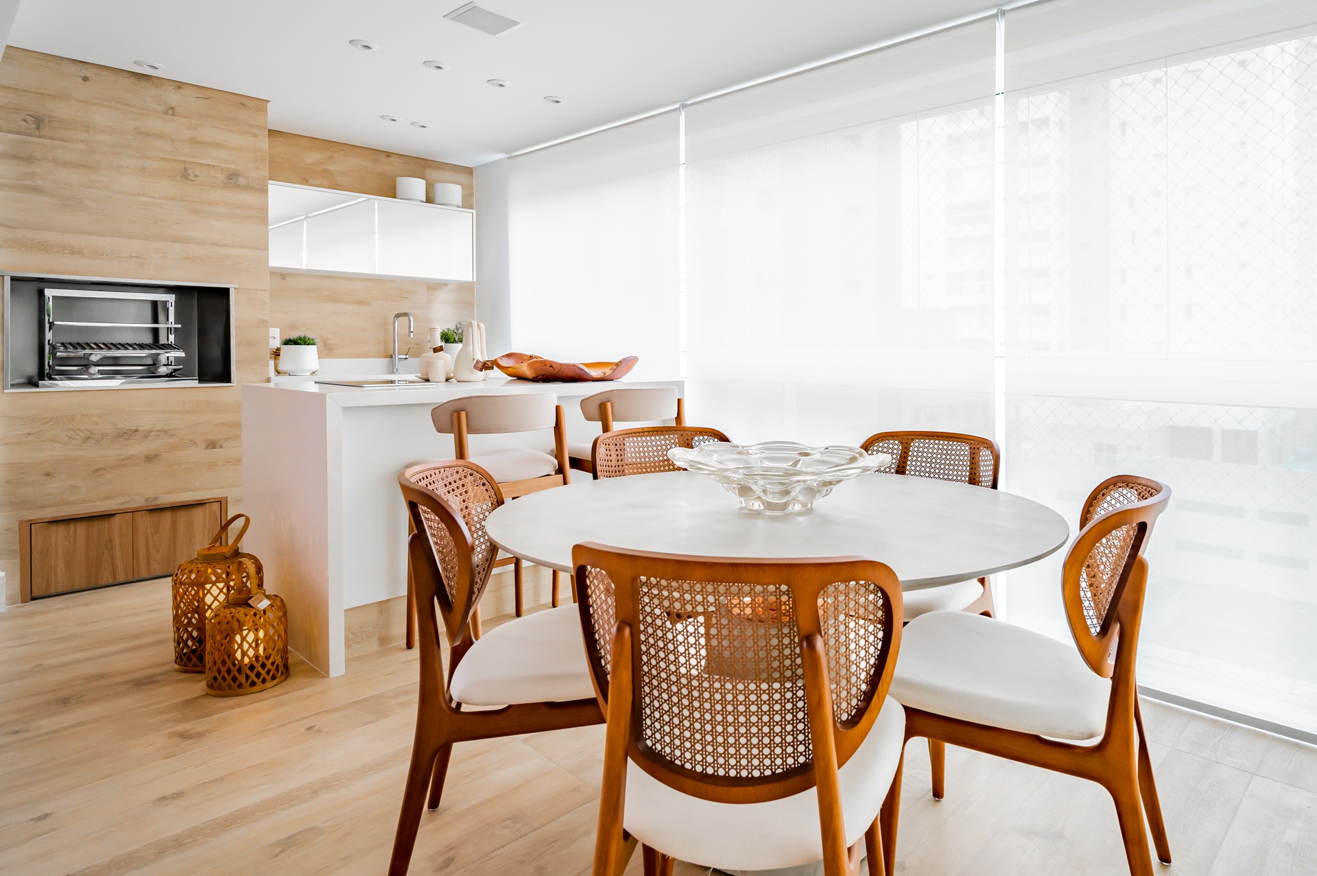 Cozinha integrada com sala de jantar, piso e parede revestidos com porcelanato que reproduz madeira, mesa redonda branca e cadeiras de madeira com encosto em palhinha
