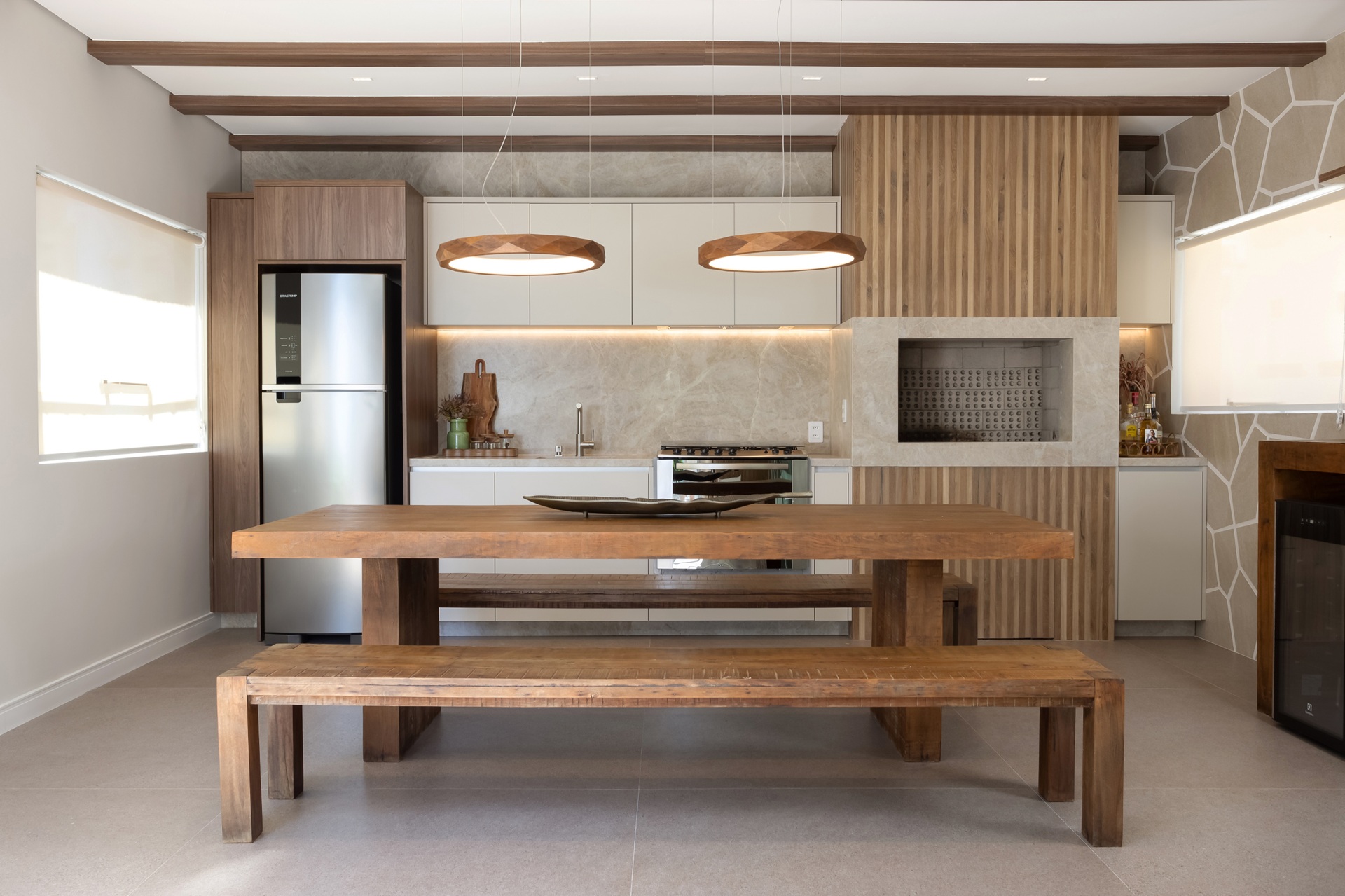 Cozinha com mesa e bancos de madeira, pendentes circulares em tom amadeirado, armários claros e churrasqueira revestida em pedra