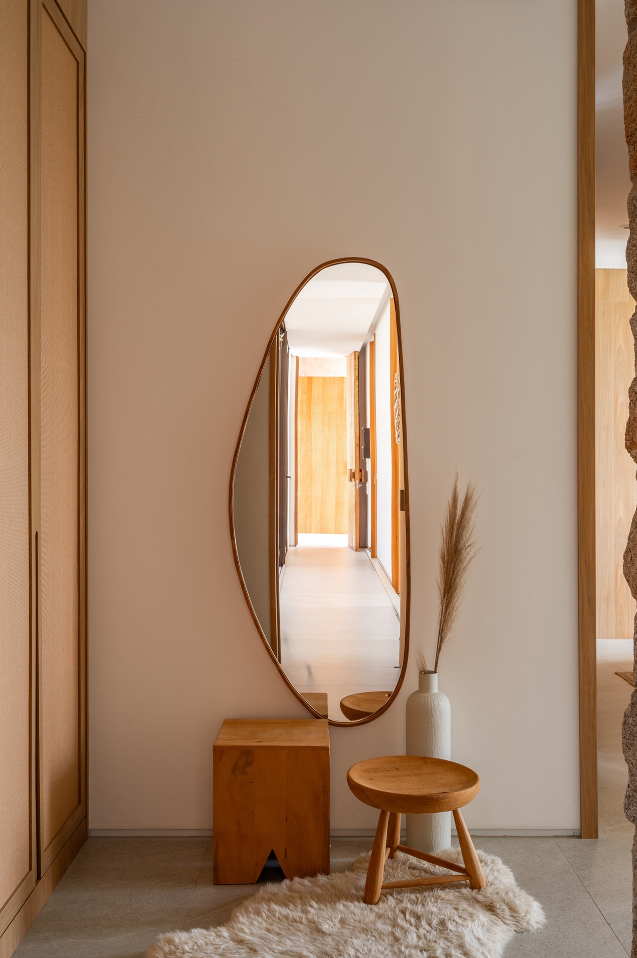 Hall com espelho orgânico de moldura em madeira, banqueta e banco baixos de madeira, tapete felpudo claro e vaso branco com ramo seco decorativo