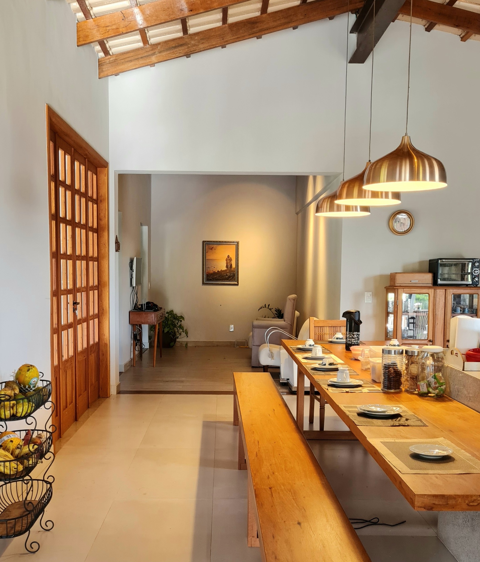 Sala de jantar com mesa retangular de madeira, bancos longos, luminárias pendentes metálicas e decoração simples com frutas e utensílios
