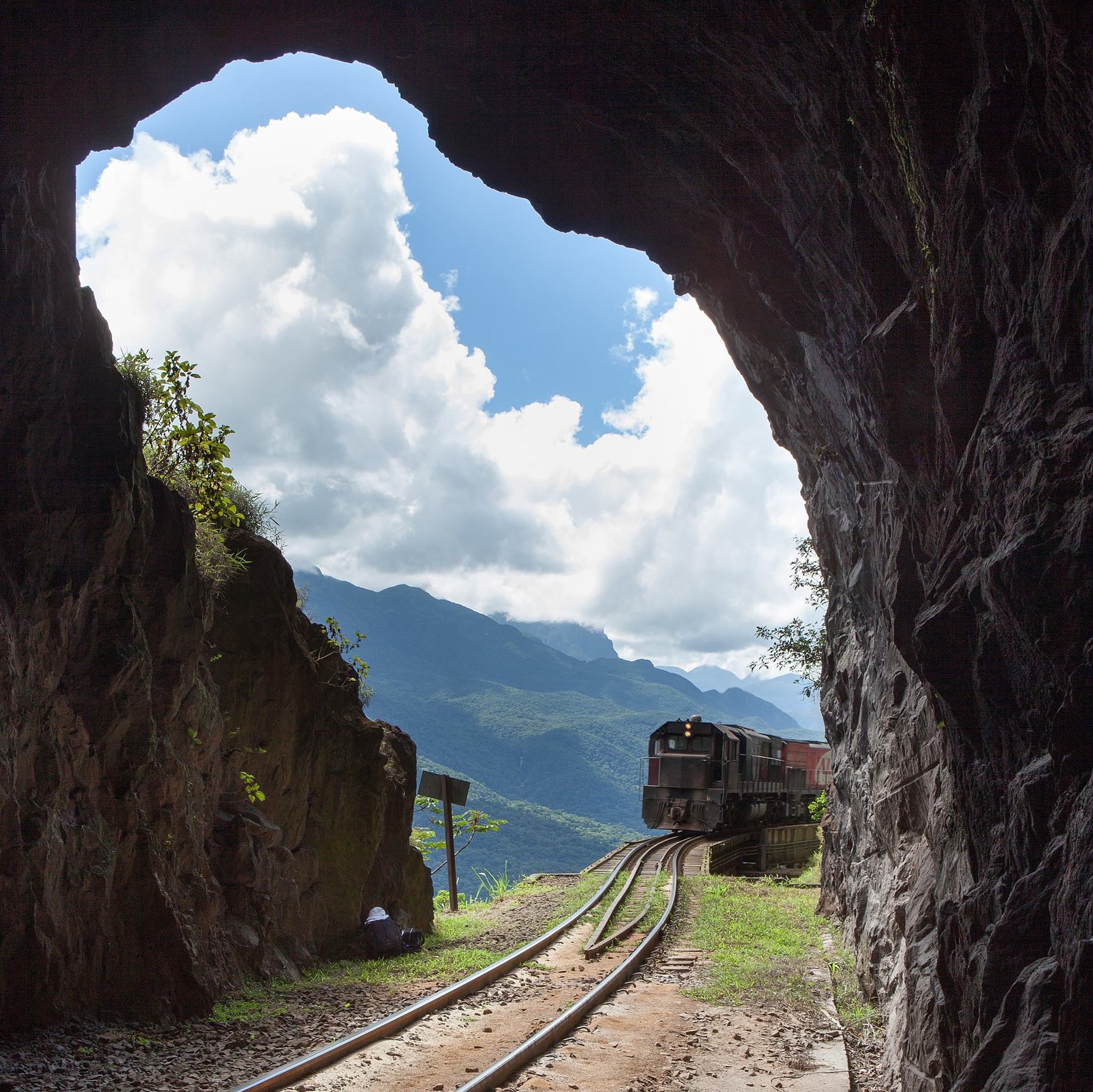 irmaos rebouças Fotografia de um trem cargueiro surgindo de um túnel na Estrada de Ferro Curitiba–Paranaguá, no Paraná. A composição enquadra a locomotiva avançando sobre os trilhos com montanhas e nuvens ao fundo, em meio à vegetação da Serra do Mar