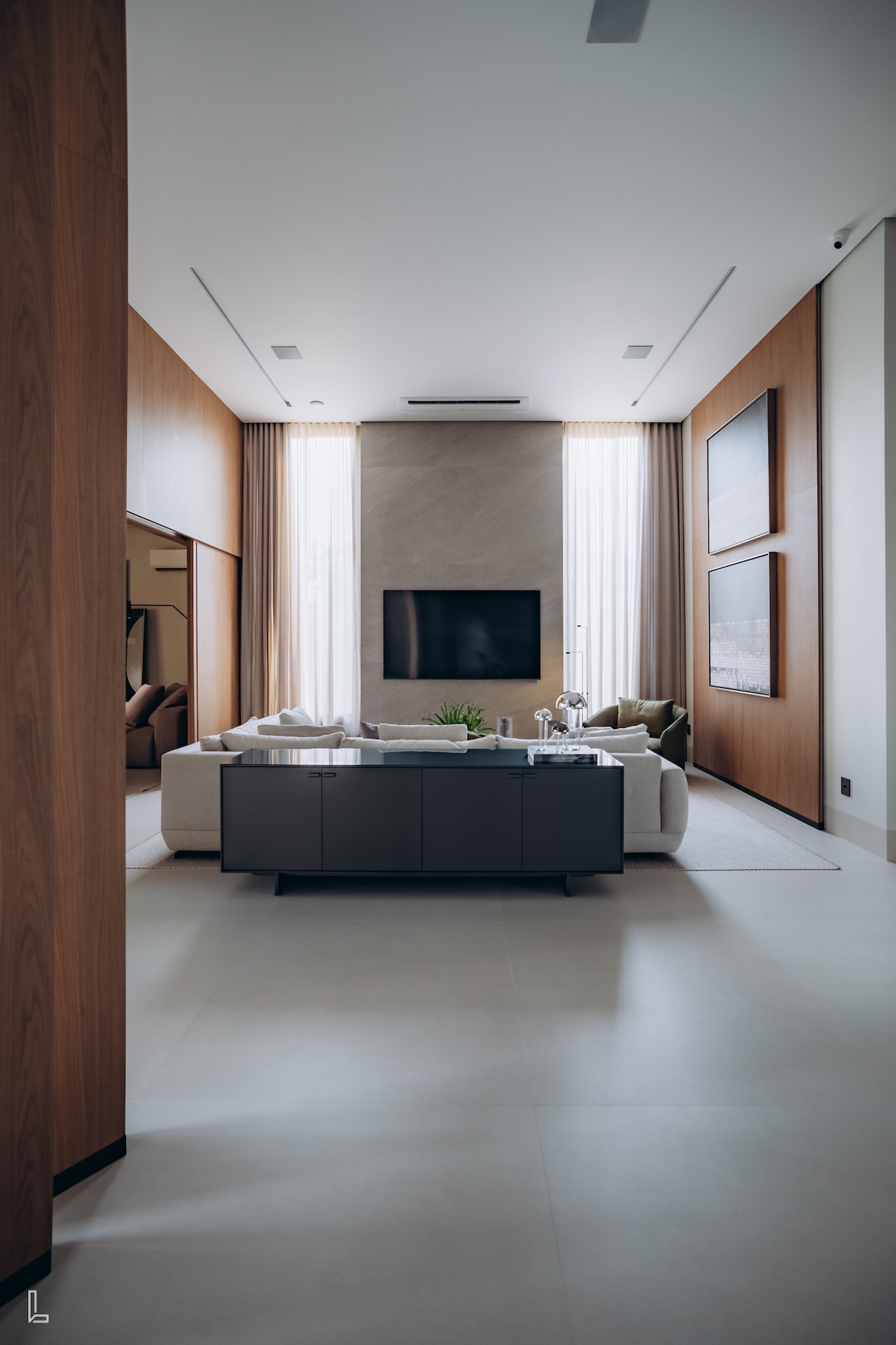 Sala ampla com televisão na parede ao lado de duas janela com cortinas translúcidas