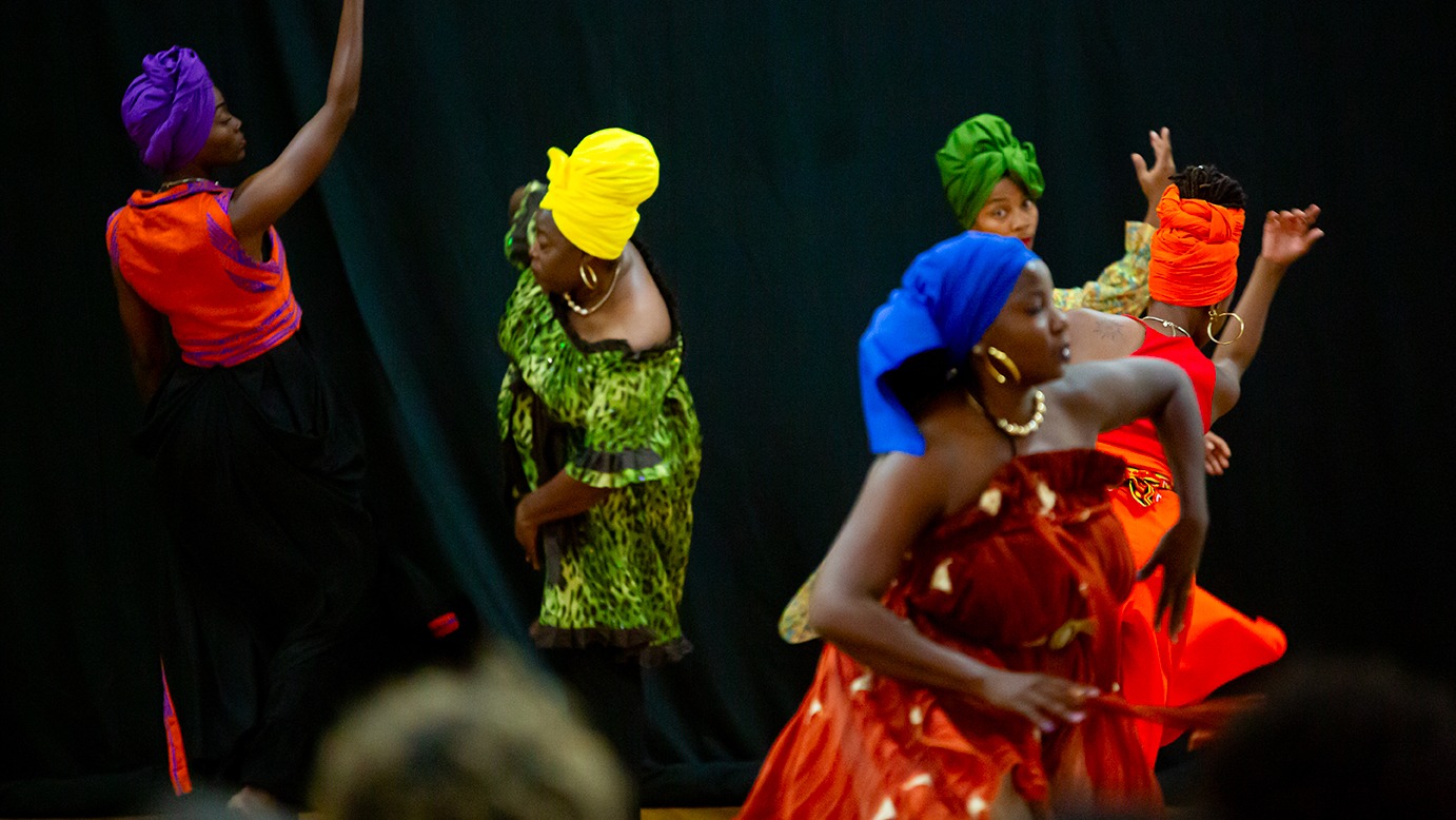 Grupo de mulheres negras dança em frente a um fundo escuro, usando roupas coloridas e turbantes vibrantes em tons de roxo, verde, amarelo, azul e laranja